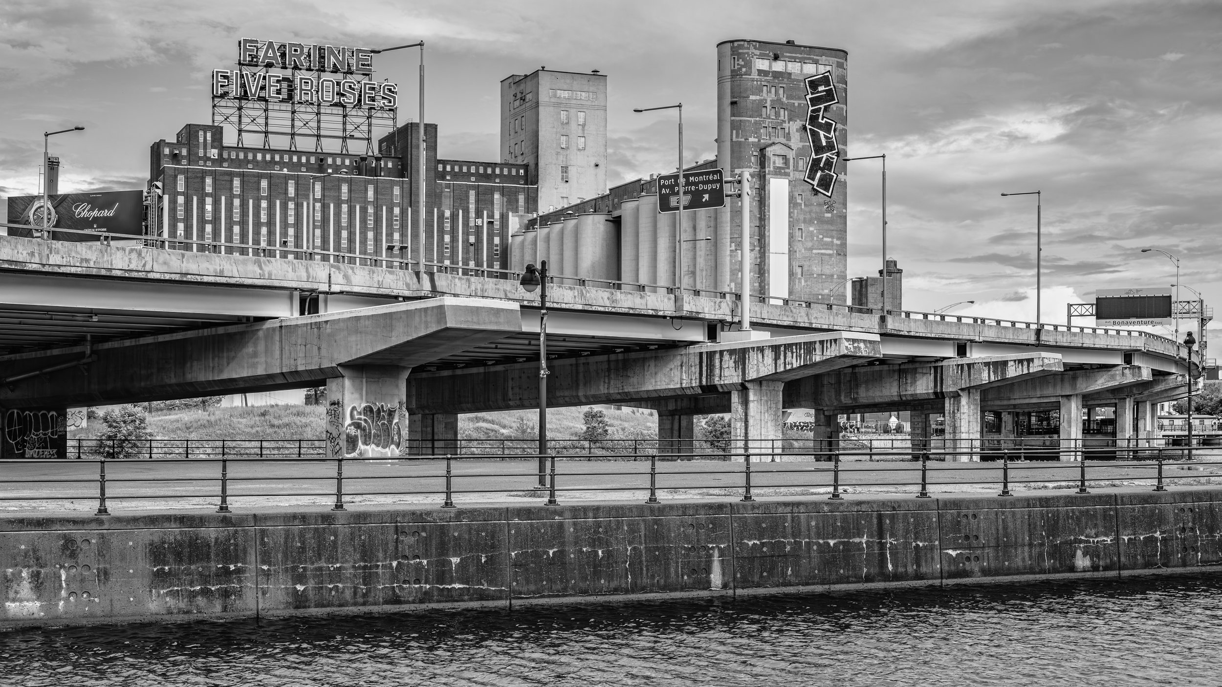 Black and white photo of an urban area with a bridge over water, graffiti on the bridge support, tall buildings in the background, and street signs indicating directions to Montreal and Pierre-Dupuy Avenue.