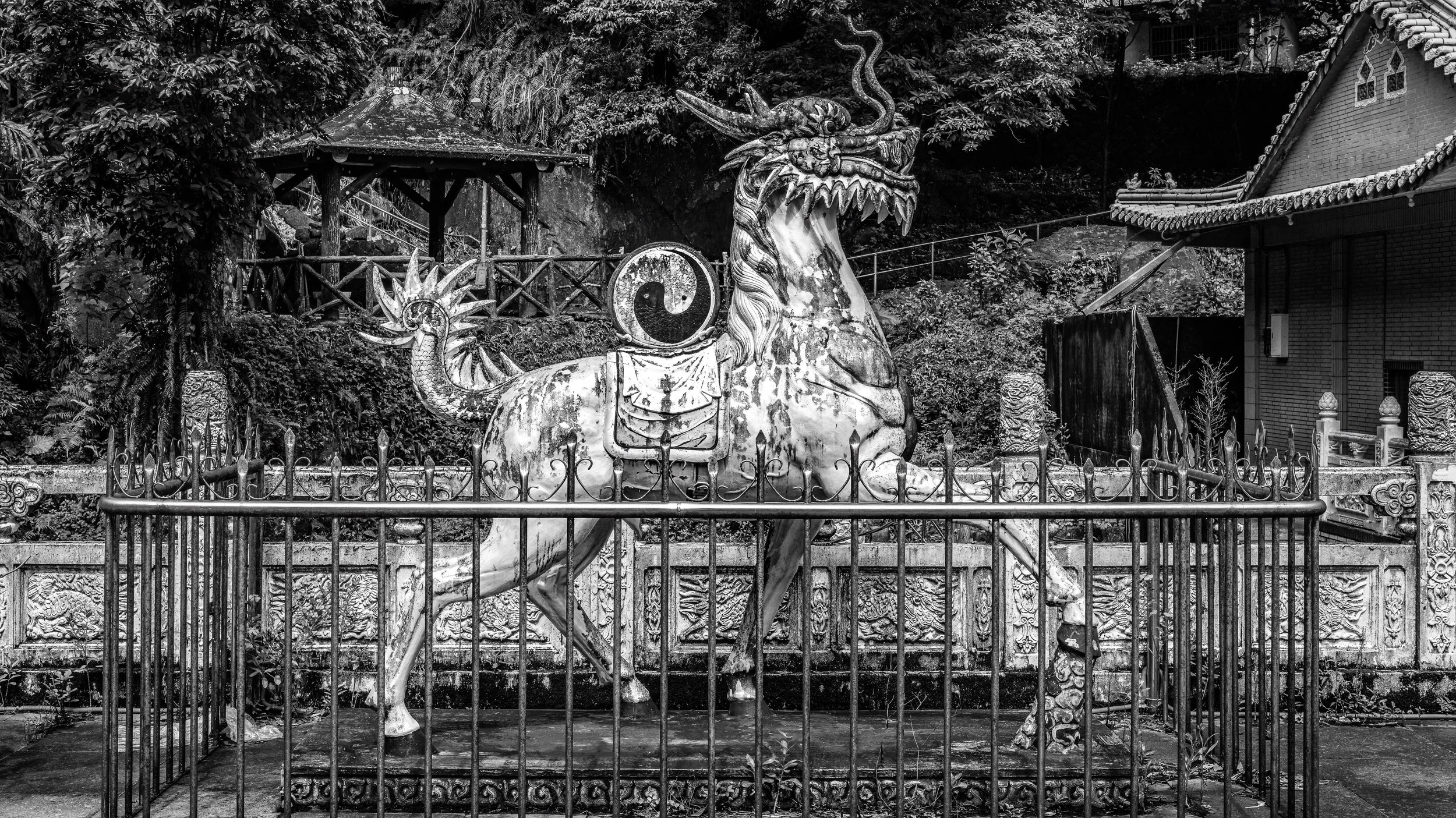 A black and white photo of a Chinese dragon sculpture on a horse statue, surrounded by a metal fence, with traditional buildings and trees in the background.