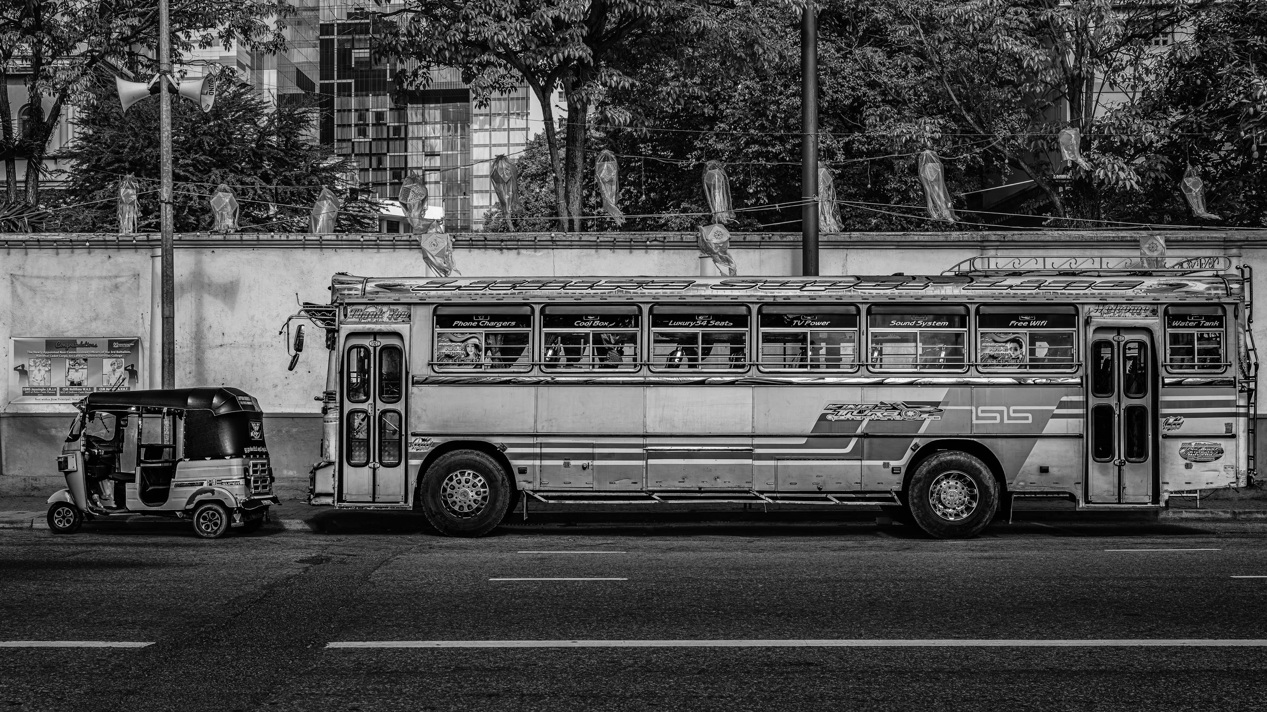 A black-and-white photo of a city street showing a small auto rickshaw and a large touring bus parked side by side. Behind the vehicles, trees and a wall with plastic bags hanging on a line can be seen, along with office buildings in the background.