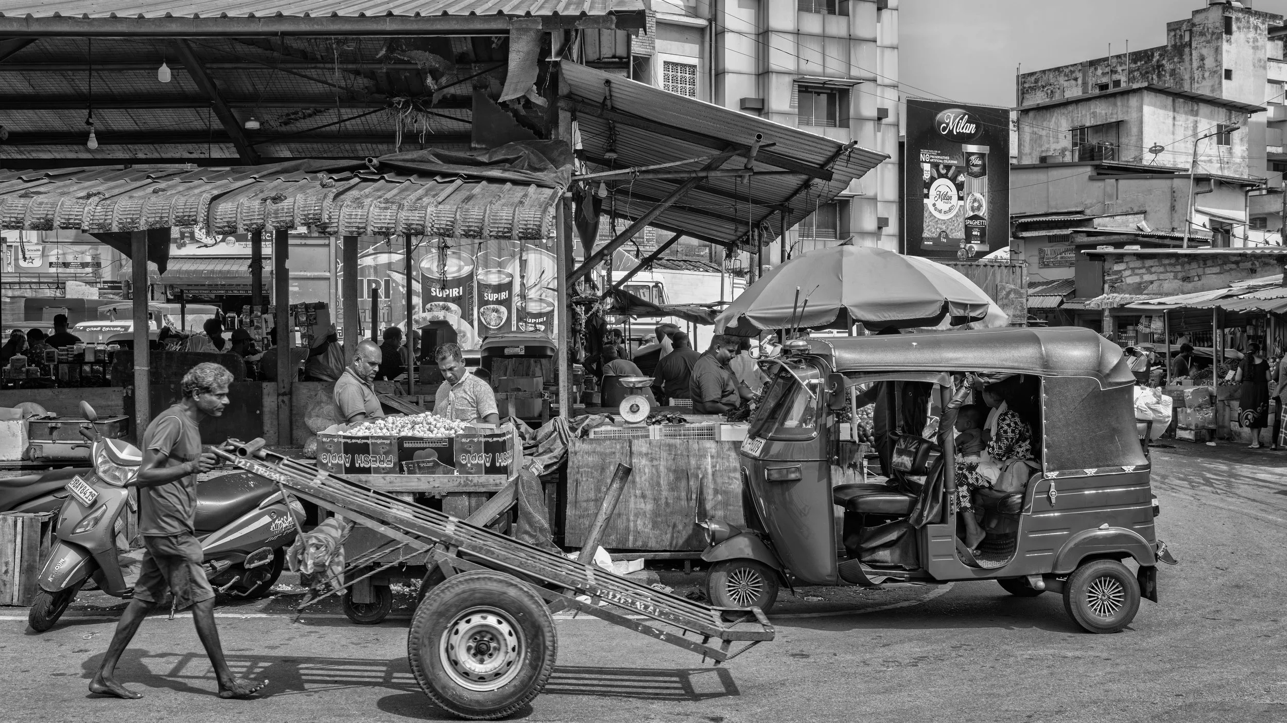 A busy outdoor market street scene in black and white with stalls selling goods, people shopping and working, a tuk-tuk, and buildings in the background.