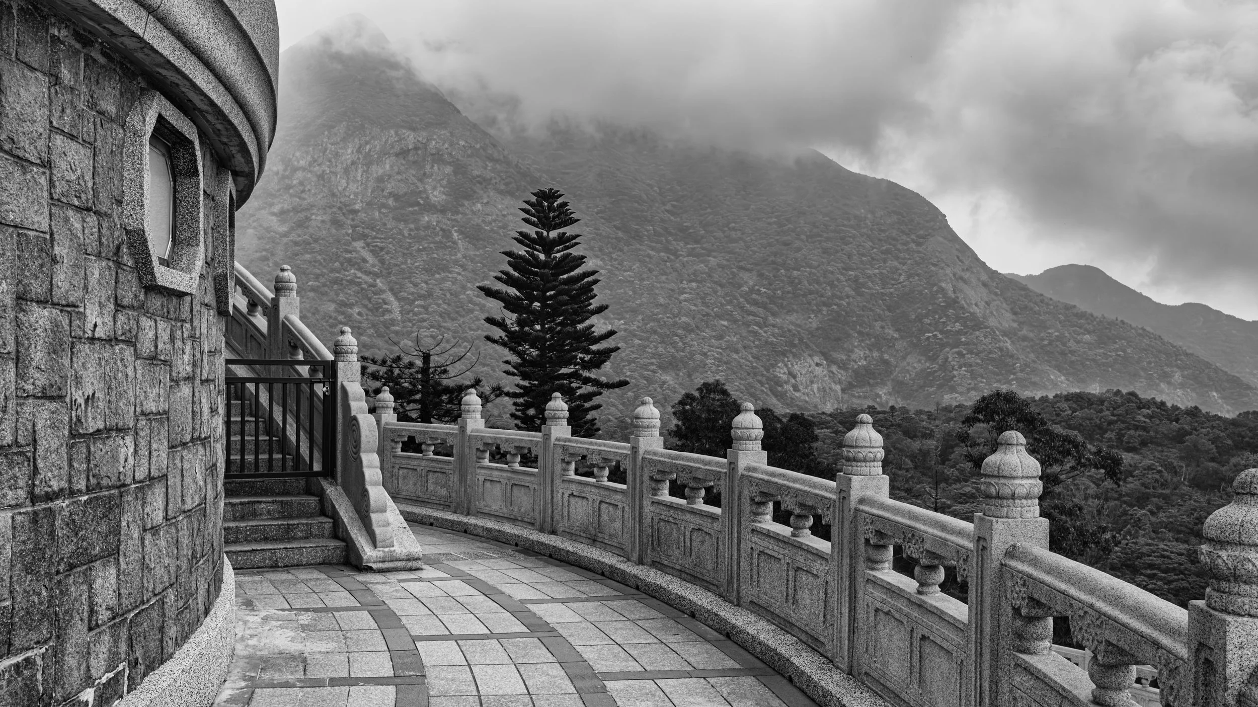 Black and white photo of a stone building with a curved wall and small window, stairs, ornate stone railing, tall pine tree, and mountains with cloudy sky in the background.