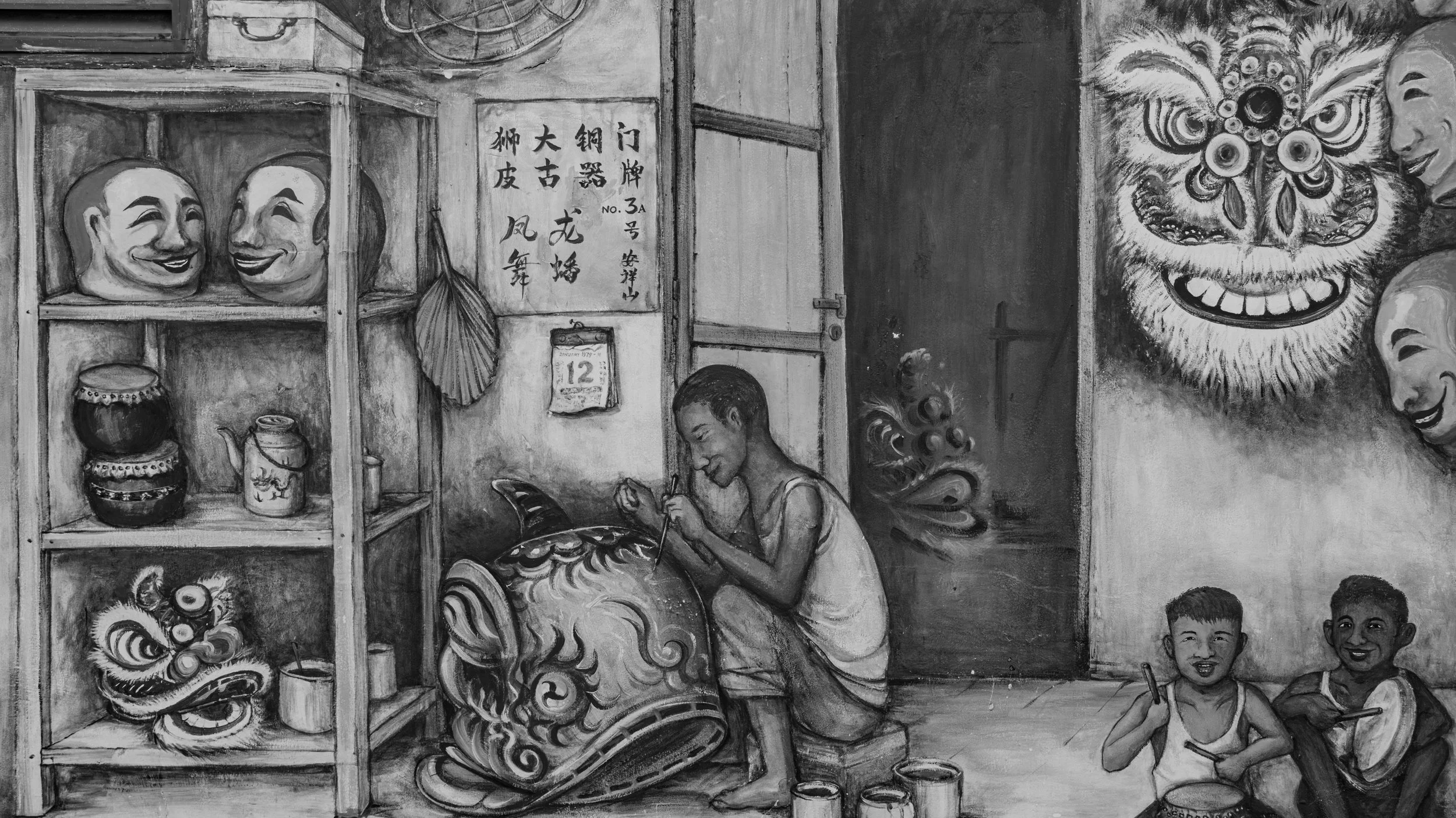 A black and white mural depicts children and traditional masks. A young boy is painting or carving a decorative item, while a boy and girl sit nearby, smiling. Shelves in the background hold masks, jars, and a fan. There is a large ornate face mask o