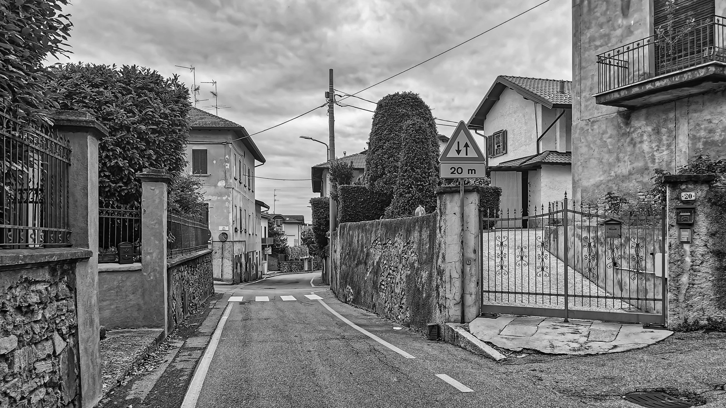 A black and white photo of a narrow urban street with houses, fences, and gates on both sides. There is a road sign indicating a two-way street 20 meters ahead, and overhead power lines against a cloudy sky.