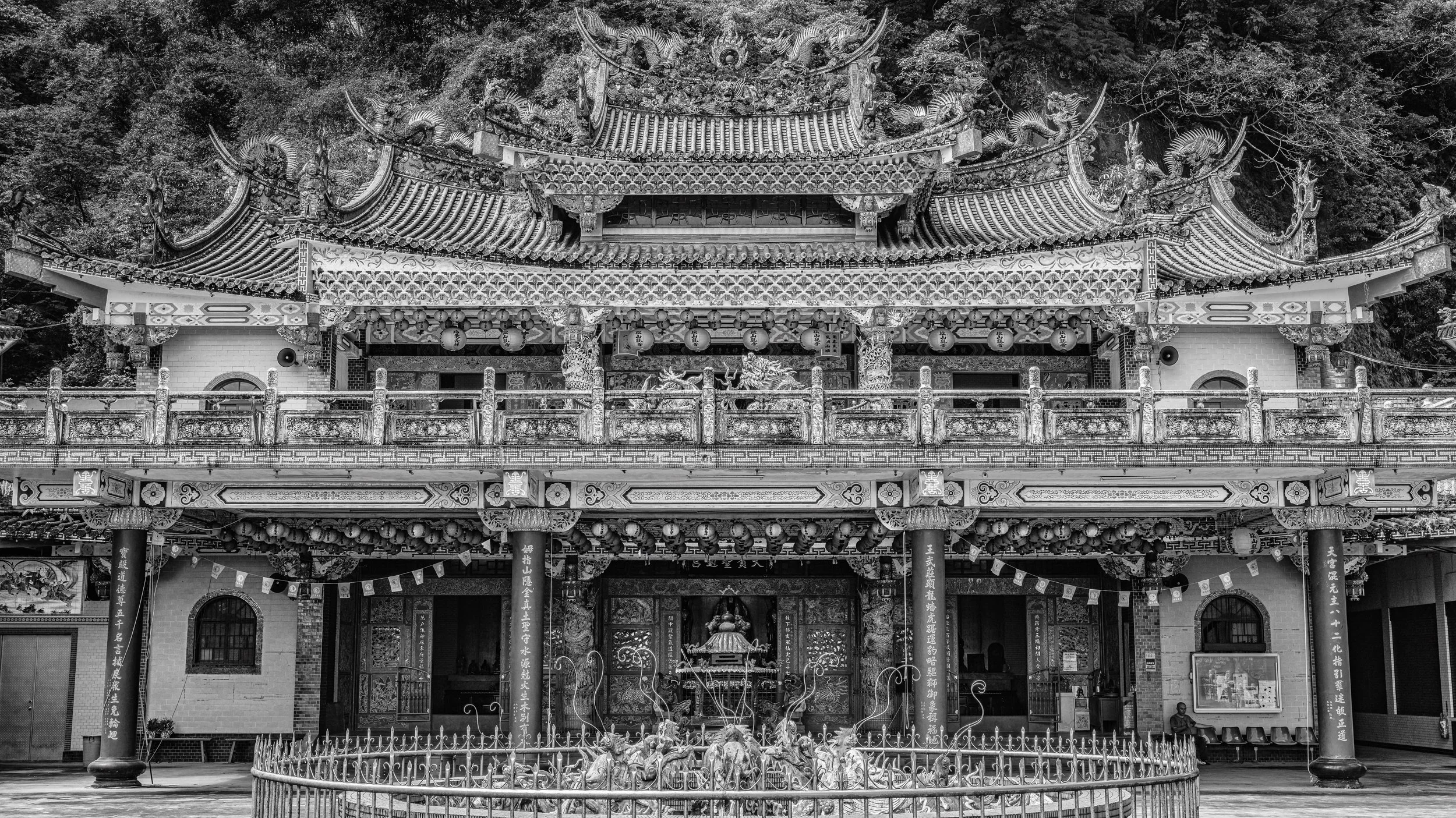 Black and white photo of an ornate Chinese temple with detailed carvings, statues, hanging lanterns, and a decorative roof, set against a backdrop of trees.