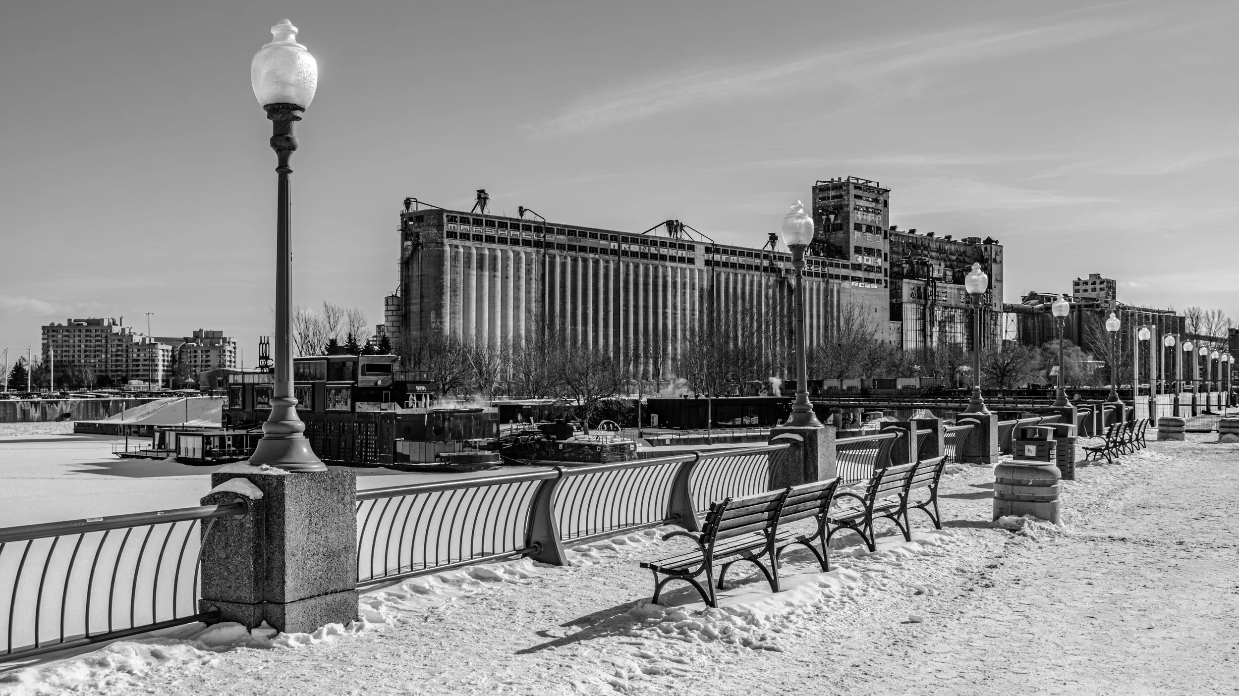 A black-and-white photo of a snow-covered waterfront park with benches, lampposts, and a historic industrial building in the background, possibly a grain elevator or factory.