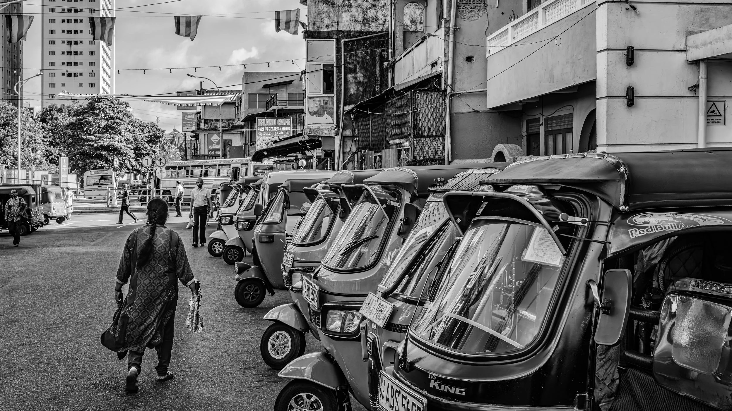 Street scene in an urban area with multiple auto rickshaws parked on the side, pedestrians walking, and buildings in the background. Black and white photo.