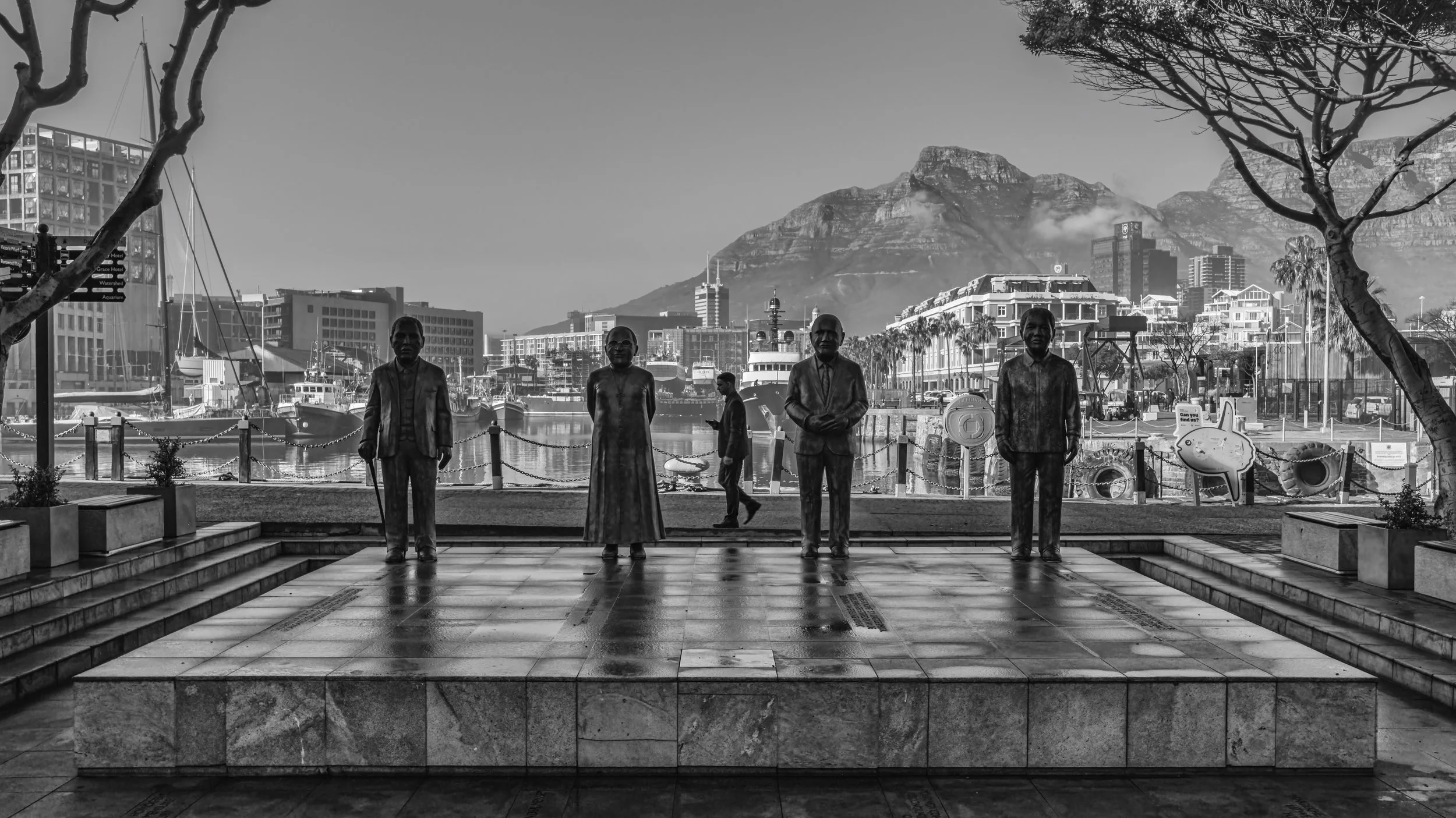 Black and white photo of a waterfront scene with mountains in the background, several statues of people in formal attire in the foreground, and boats docked at the marina. Cape Town, South Africa.
