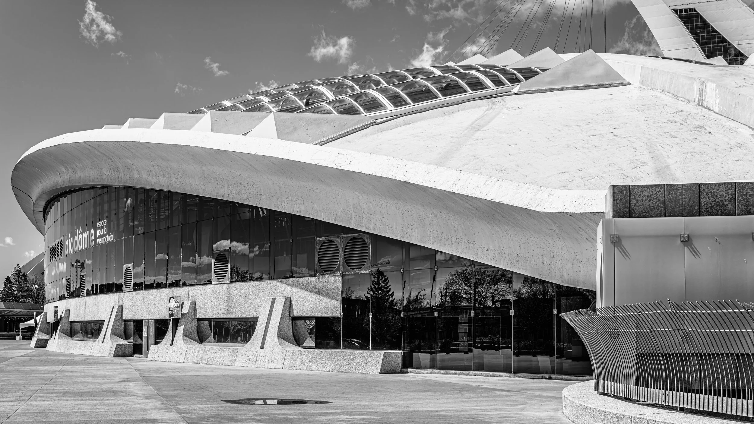 Black and white photo of a modern architectural building with curved concrete and glass design, featuring a sloped roof with glass panels and a large circular glass window at the front, with a clear sky and few clouds in the background.