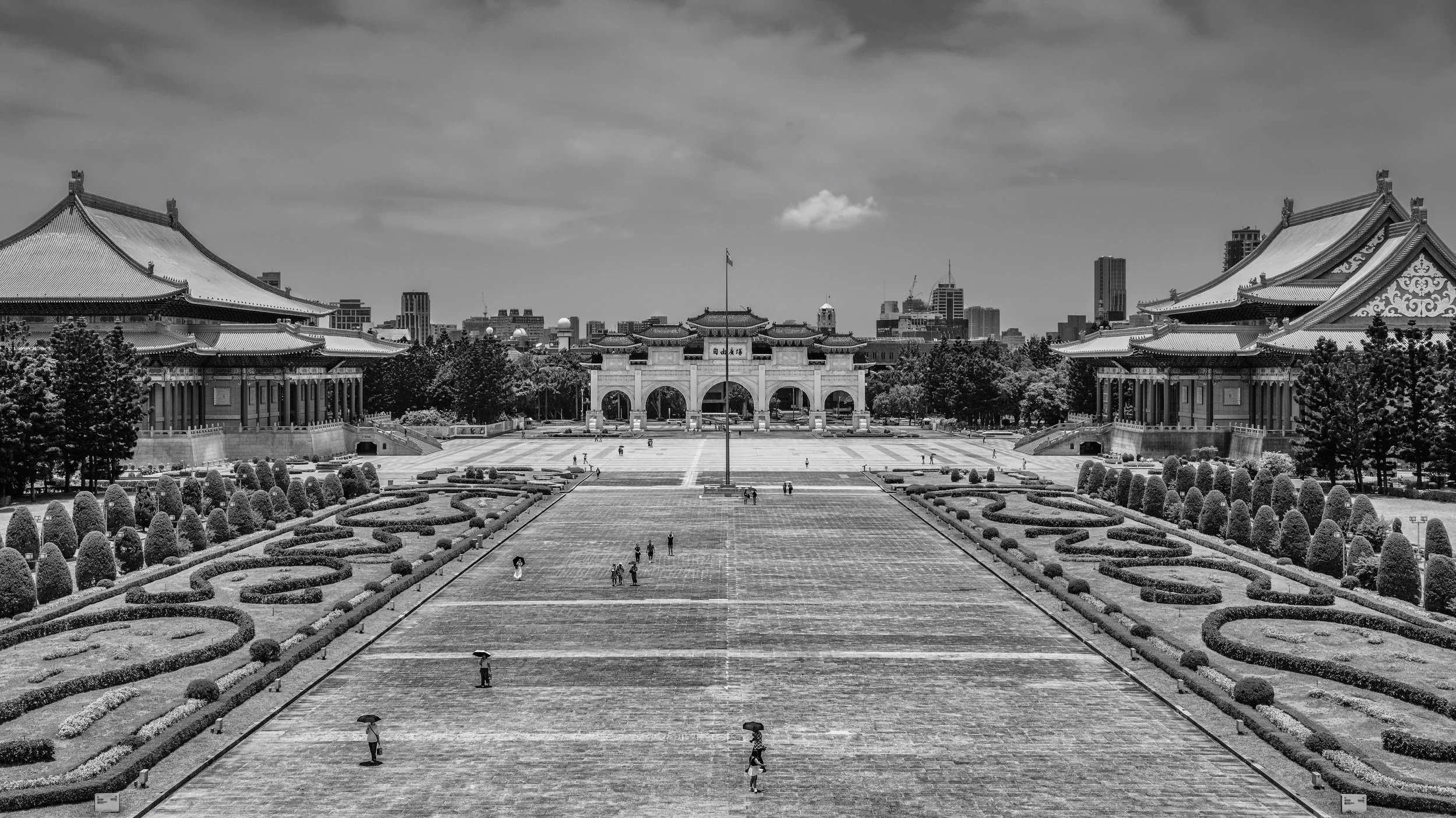 A large traditional Asian-style temple complex with ornate rooftops and decorative details, surrounded by a well-maintained garden with manicured bushes and trees, set against a modern city skyline under a partly cloudy sky.