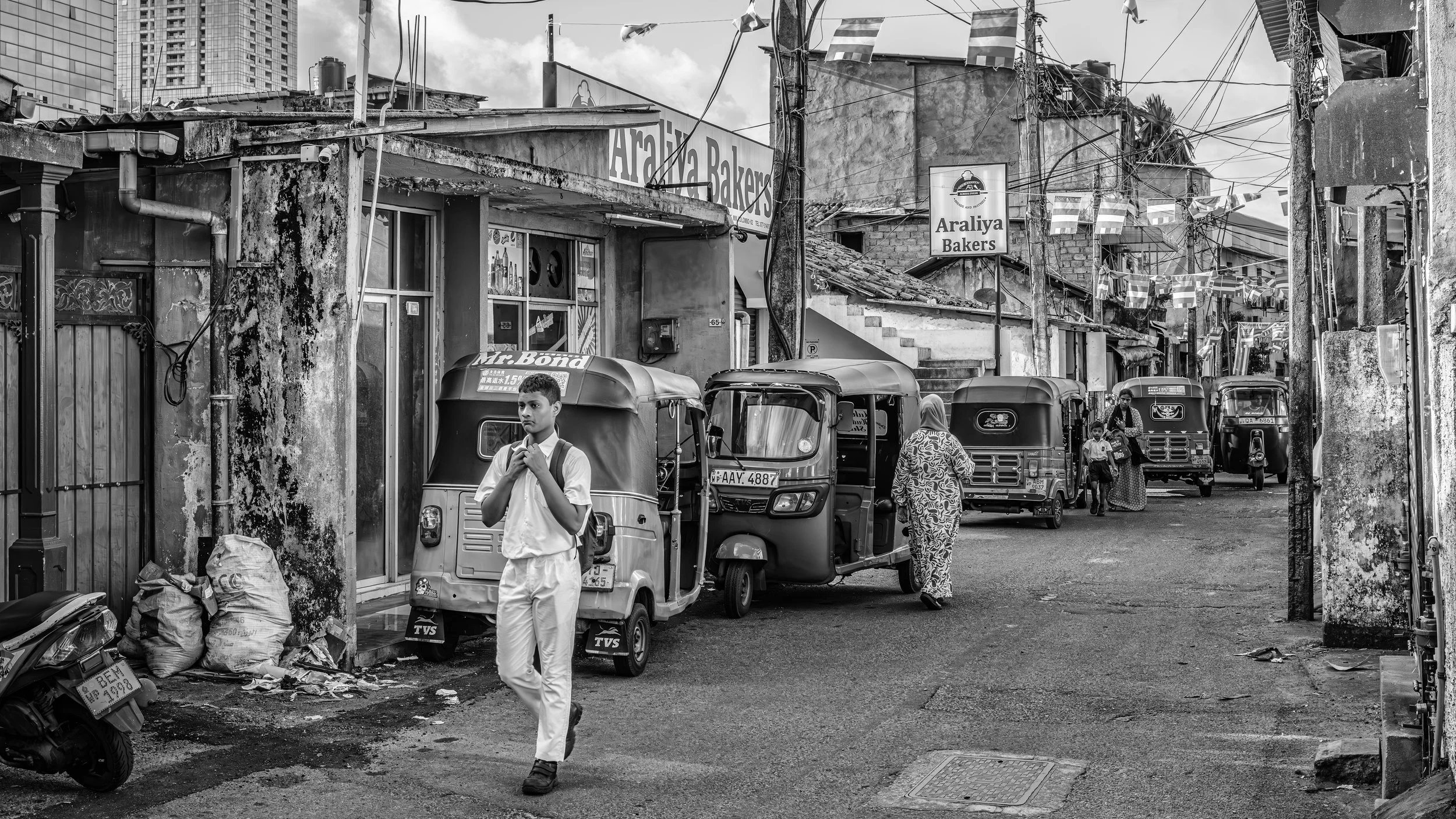 Street scene in an urban area with people and auto rickshaws, with buildings, power lines, and signs for 'Araliya Bakers' in the background, in black and white.
