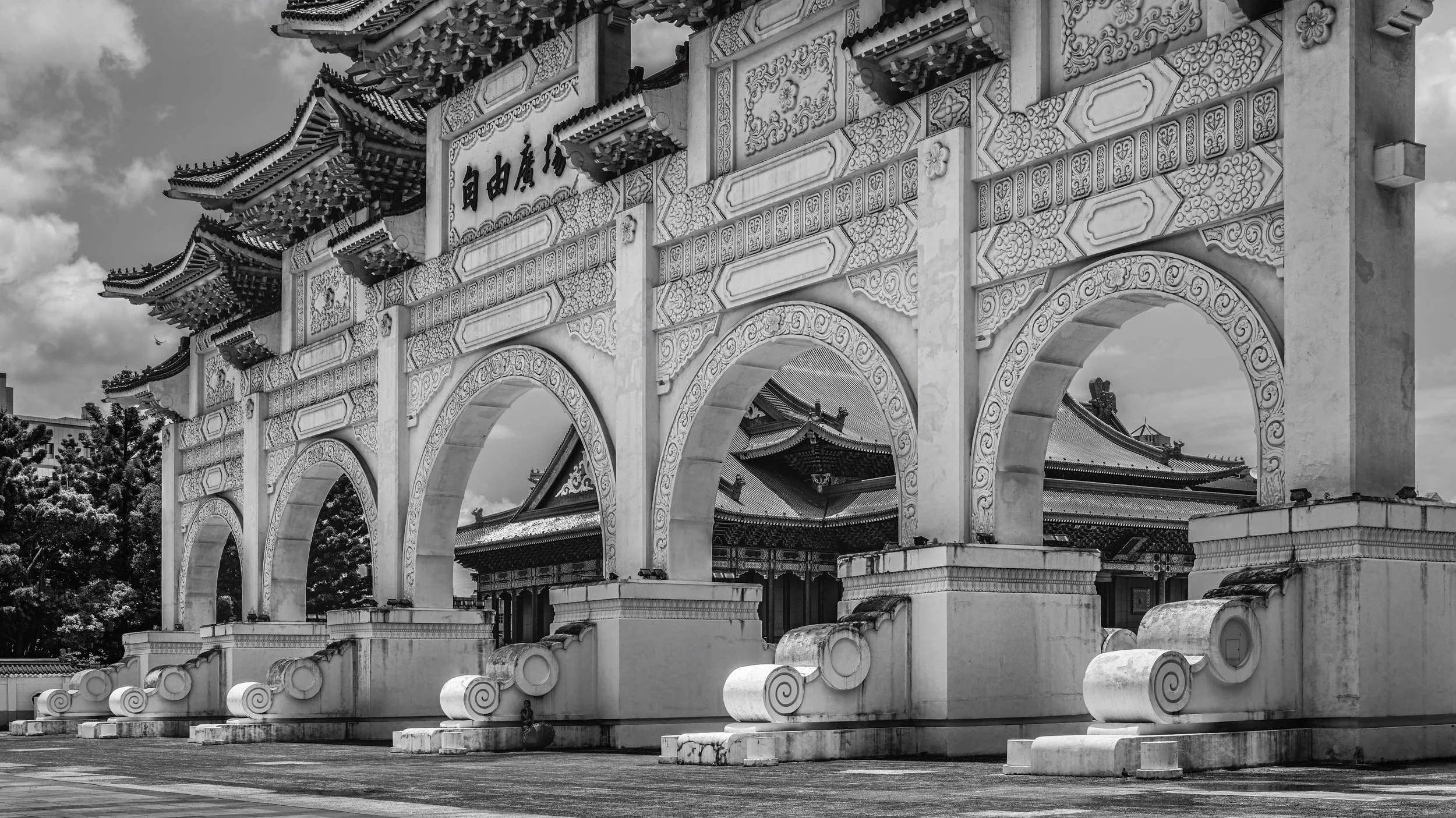 Black and white photo of a traditional Asian temple gate with decorative roof and ornate carvings, with a building visible through the arches. Taipei, Taiwan.