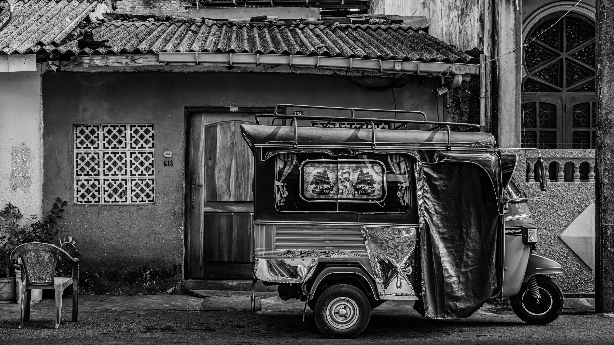 A black and white photo of an outdoor scene featuring a colorful truck parked in front of an old building. The building has tiled roofing, a decorative window on the right, and a small square vent on the left. An empty chair and a small table are vis