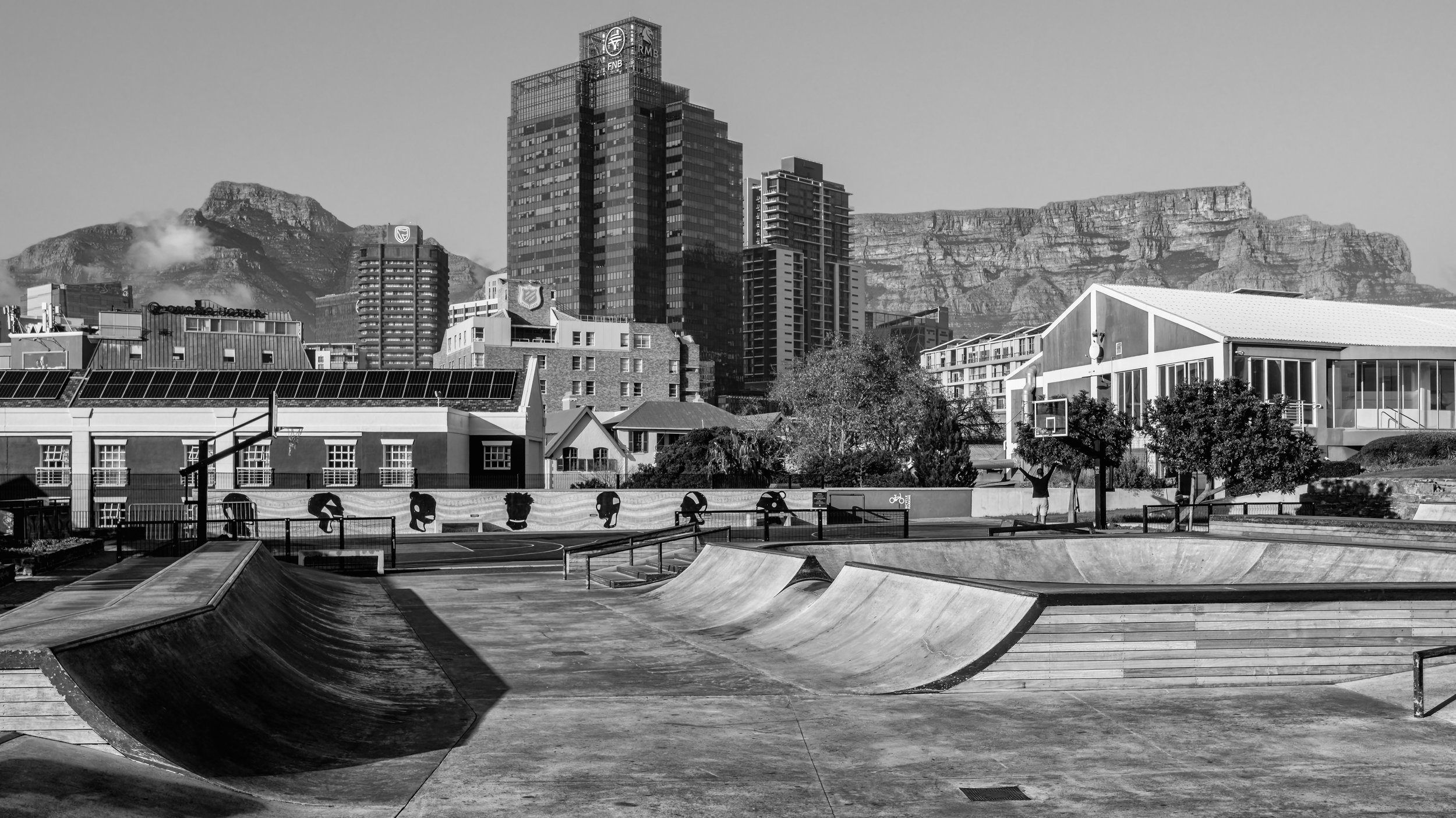 A city skyline with tall buildings and mountains in the background, viewed from a skatepark with ramps and railings in the foreground, in black and white. Cape Town, South Africa.