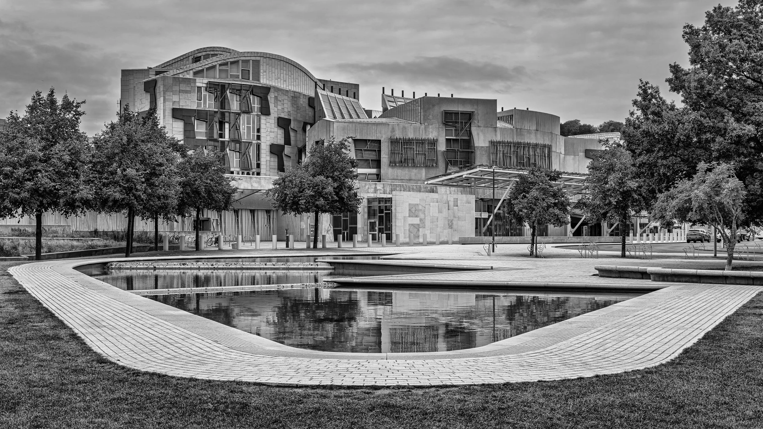 A black and white photo of a modern building with unusual architecture, trees, a small pond, and a paved walkway in the foreground.