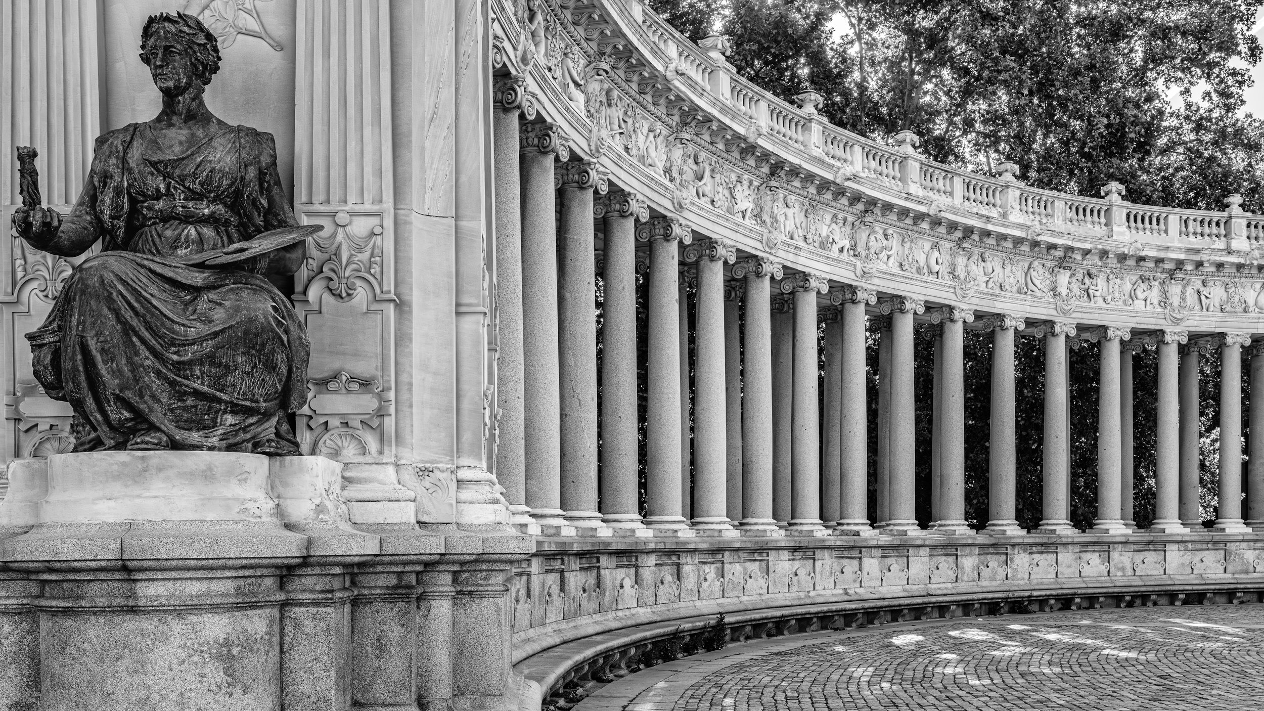 A black and white photo of a seated bronze statue of a woman holding a small object in her right hand and an open book on her lap, situated in front of a ornately decorated colonnade with columns and intricate carvings, with trees in the background.