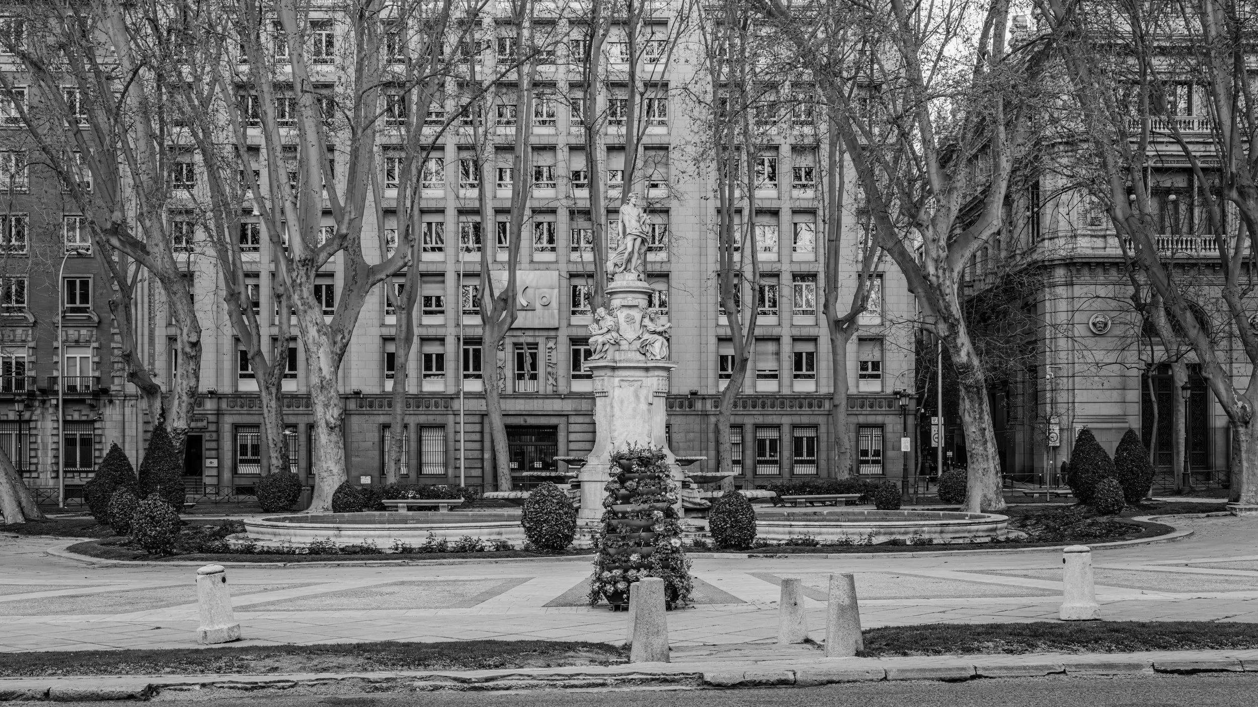 Black and white photo of a city park with a large stone statue in the center, surrounded by leafless trees and a multi-story building in the background.