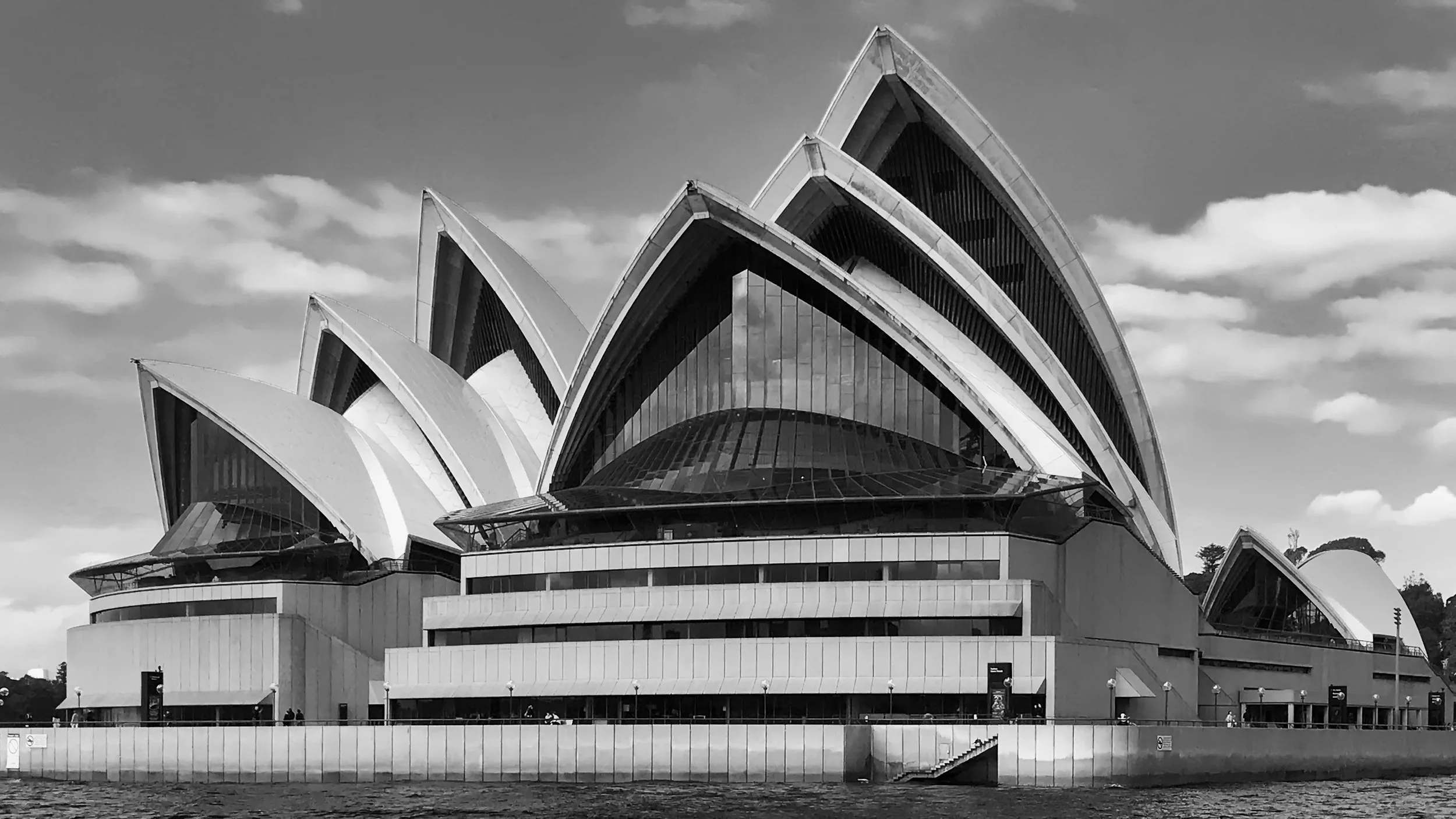 Black and white photograph of the Sydney Opera House with its distinctive sail-like roof structures, set against a partly cloudy sky.