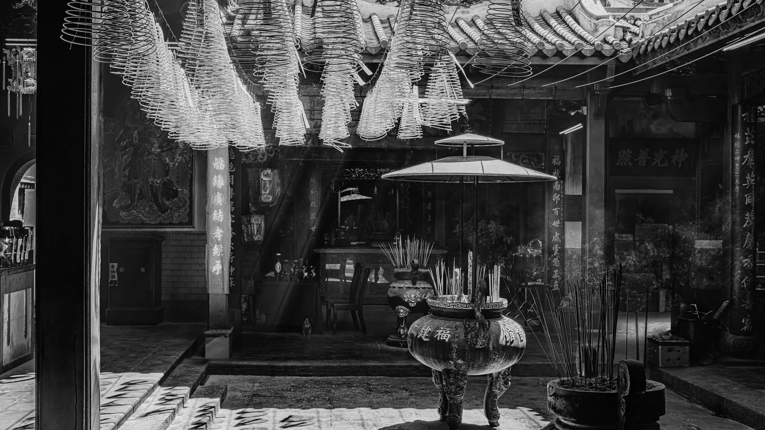 Black and white photo of traditional Asian temple interior with hanging spiral incense coils, large decorative incense burner, and ornate background decorations with Chinese characters.