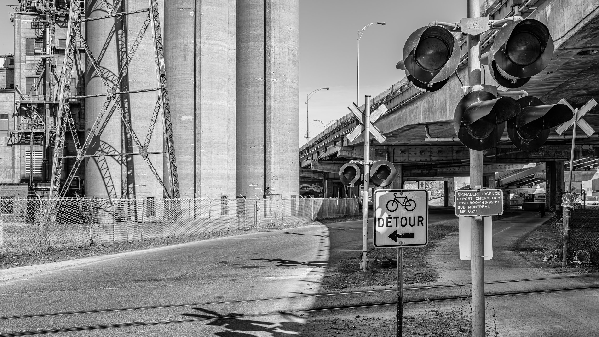 Black and white image of an industrial area with large silos, metal structures, and a railway crossing with signal lights. Signs indicating detour and emergency report in French and English are visible.