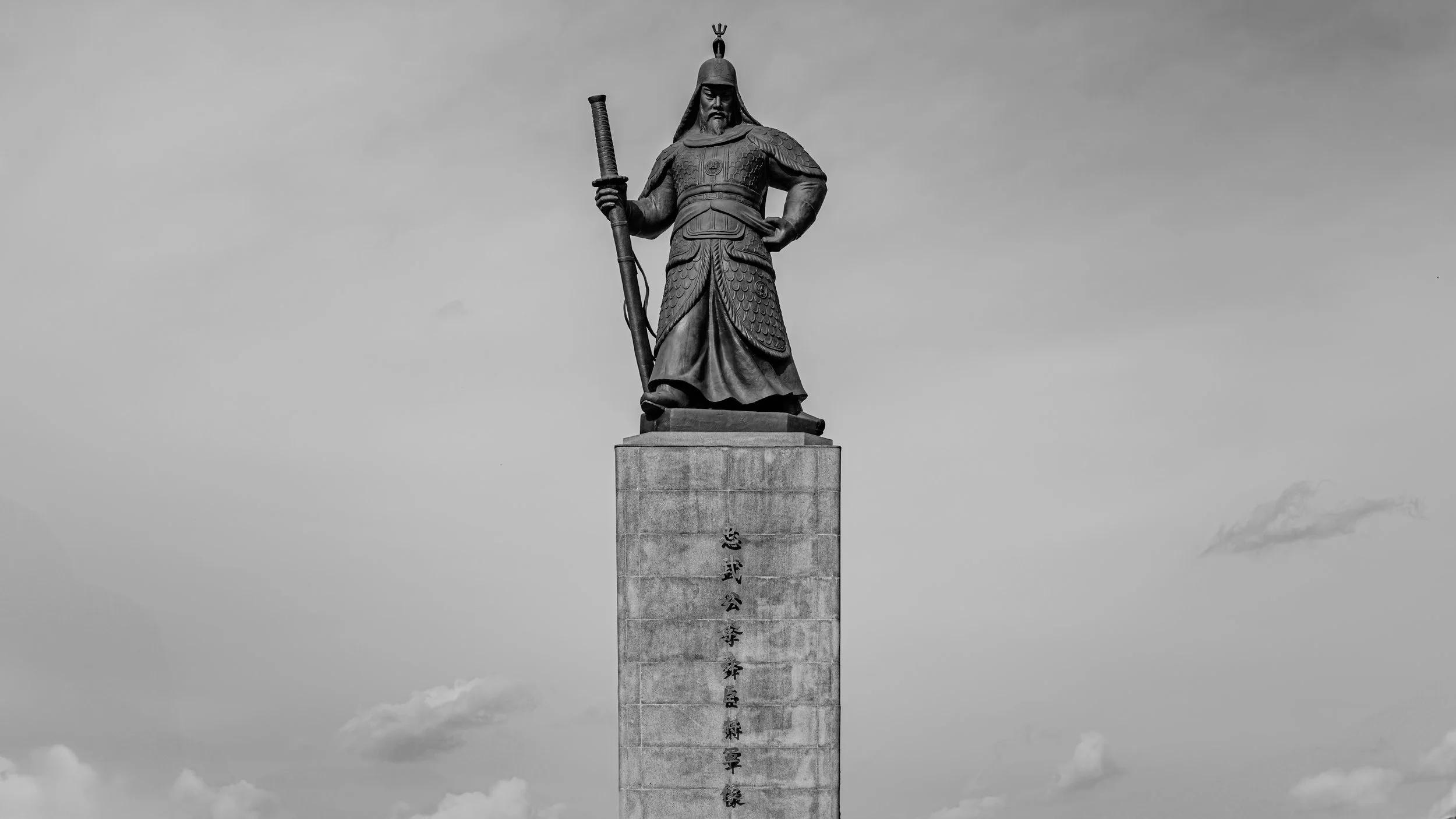 Black and white photo of a large statue of a warrior or historical figure standing on a tall pedestal with inscriptions.