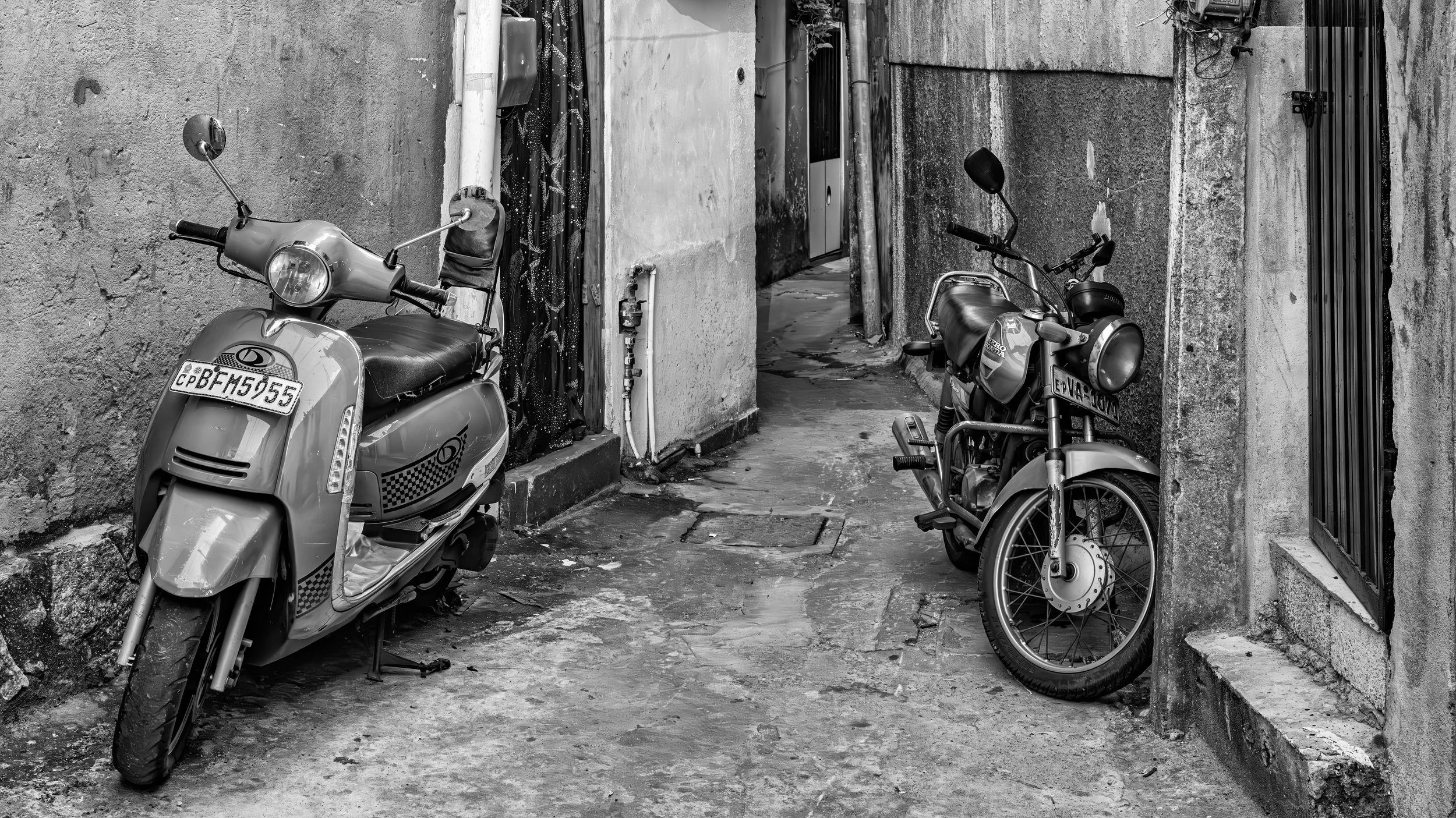 Black and white photo of two scooters parked on a narrow alleyway with worn walls and a cracked concrete ground.