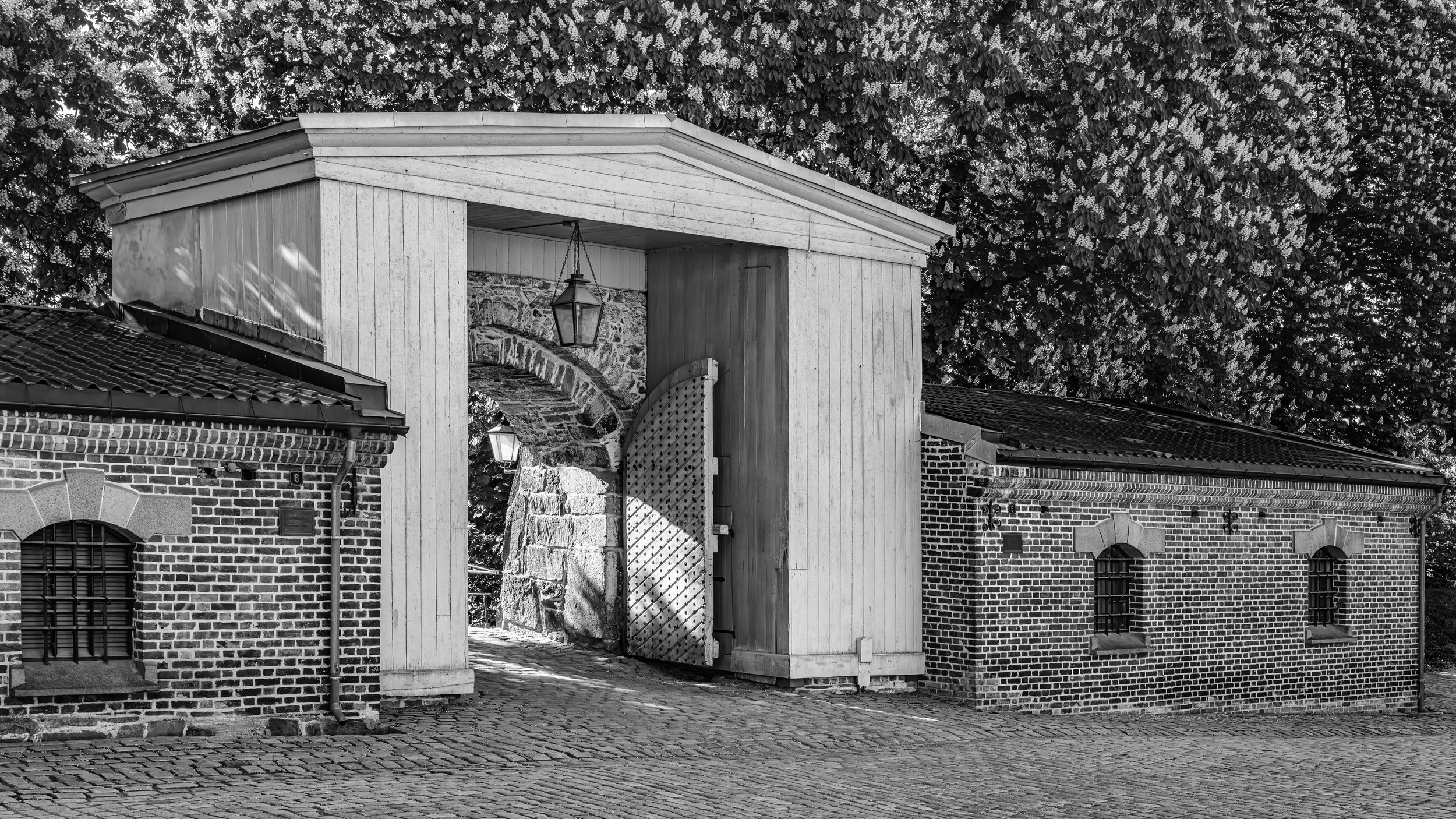 A black and white photo of a brick building with arched windows and a tiled roof, with a wooden gate and a stone archway in the background, surrounded by trees.