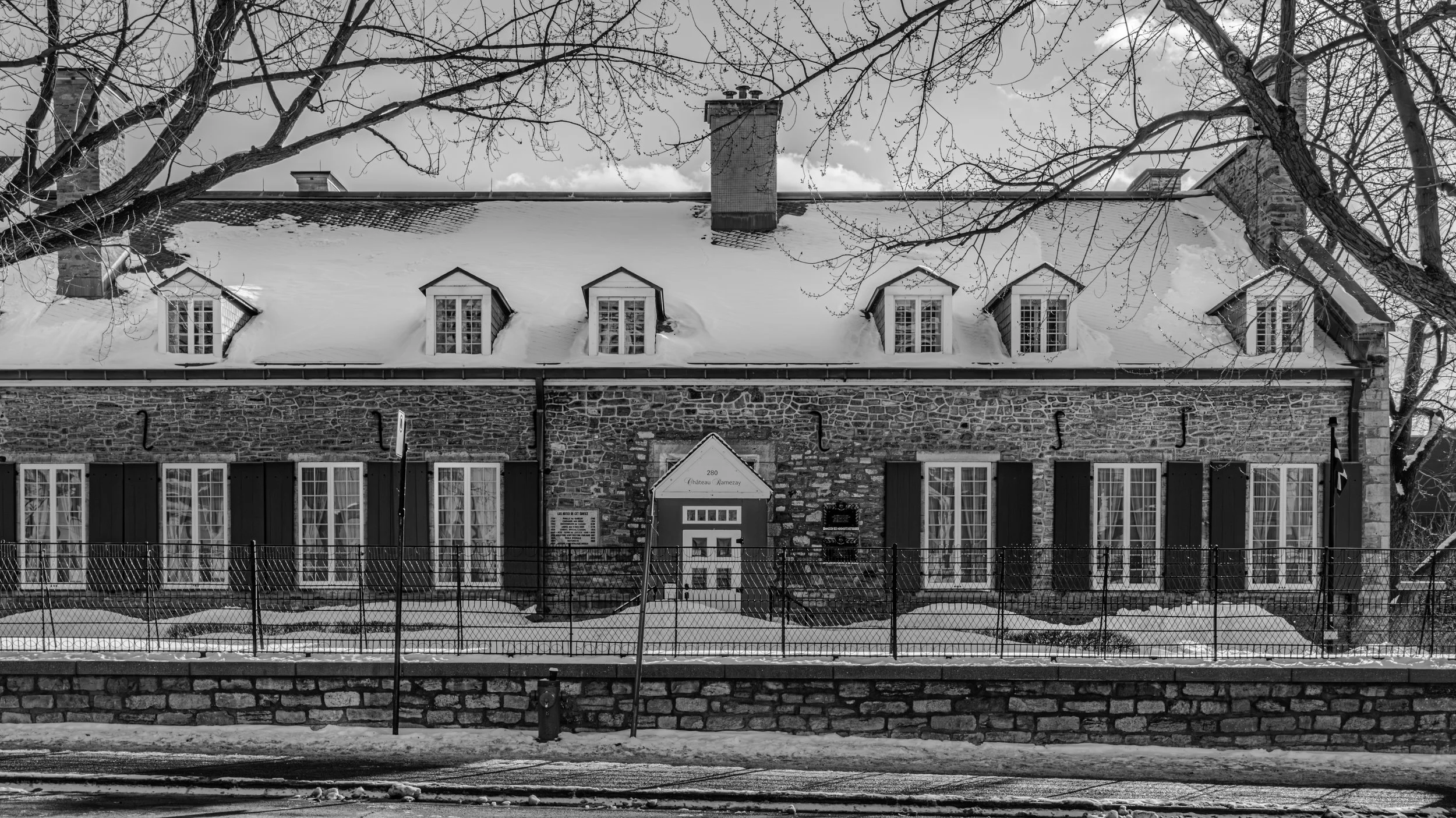 Black and white photo of a stone building with dormer windows and a snowy roof, trees without leaves in the foreground, a fence, and a street with railway tracks in front.