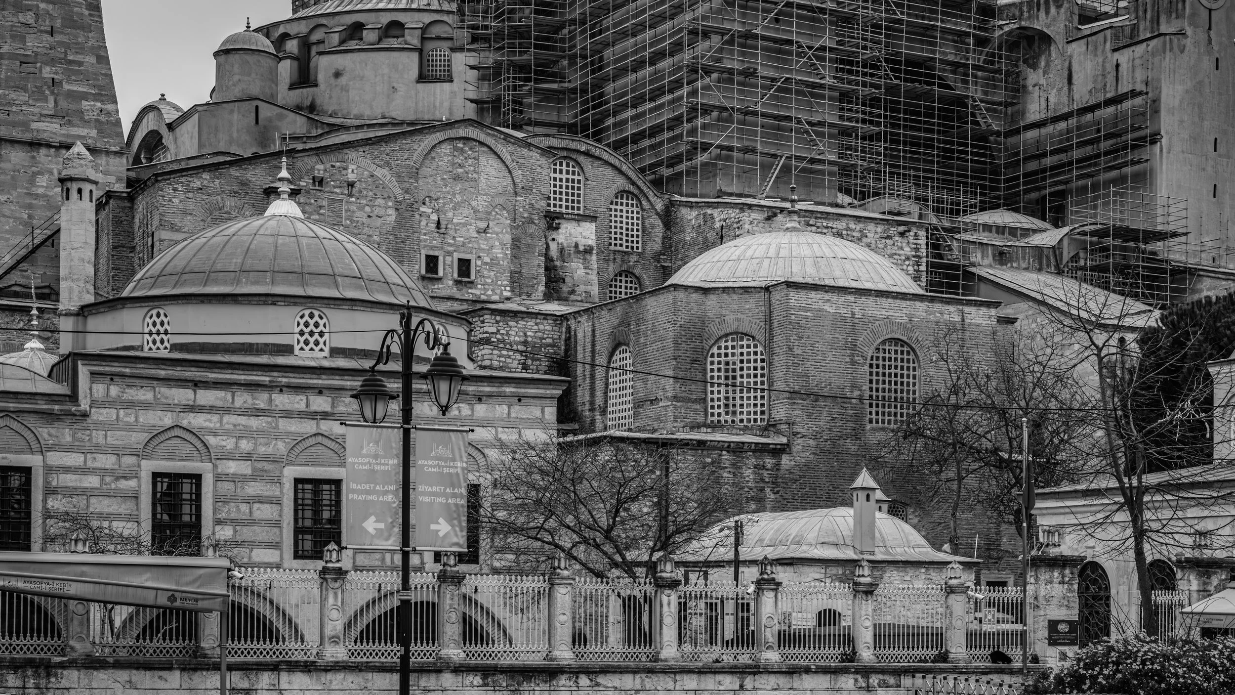 Black and white photo of an ancient mosque with scaffolding on the upper part, featuring domes, arches, and windows, with street signs and trees in the foreground.