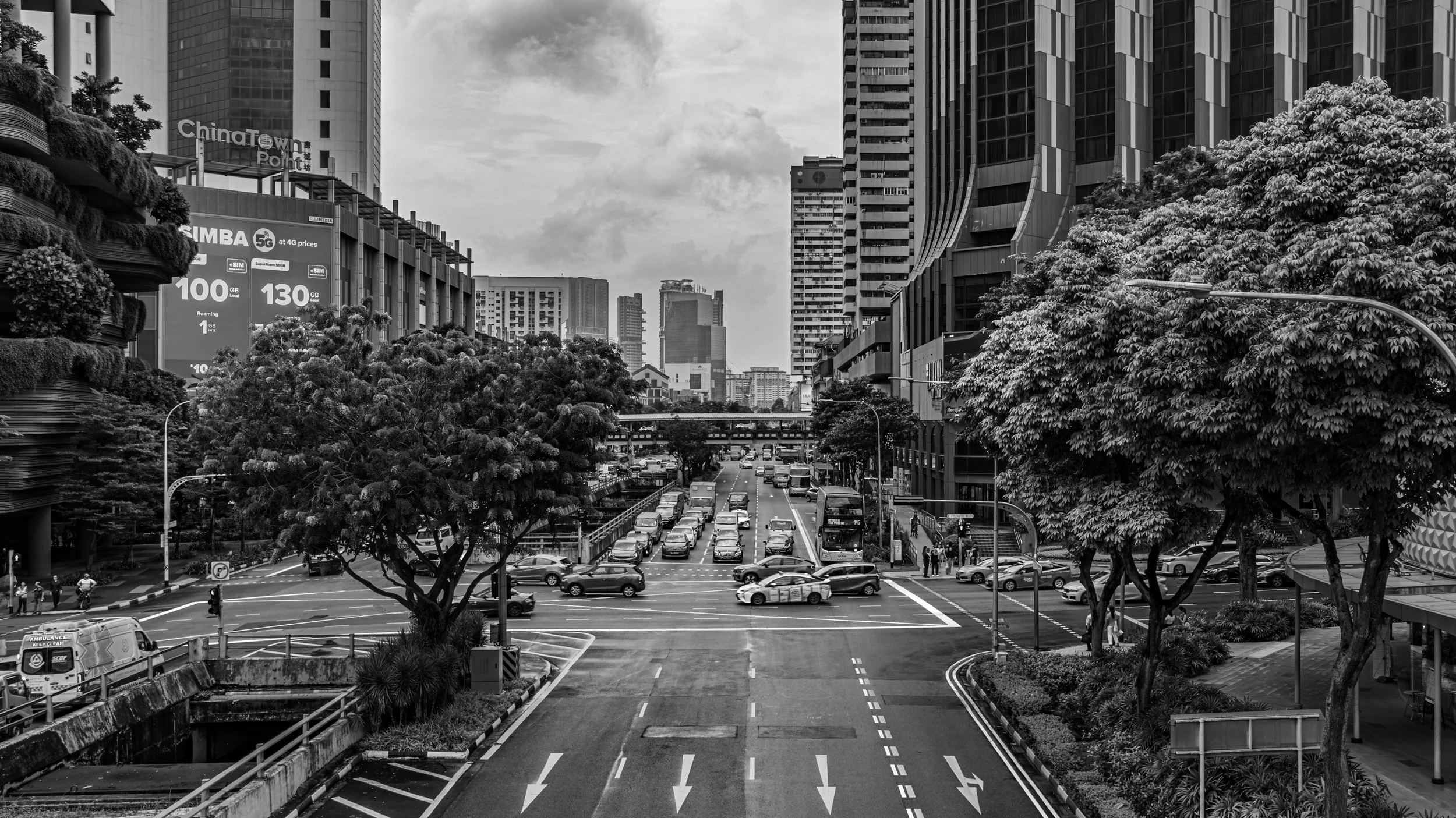 Black and white photo of a busy city street with tall modern buildings, trees, and vehicles including cars and an ambulance. Pedestrians are at crosswalks, and there are signs and billboards on the buildings.