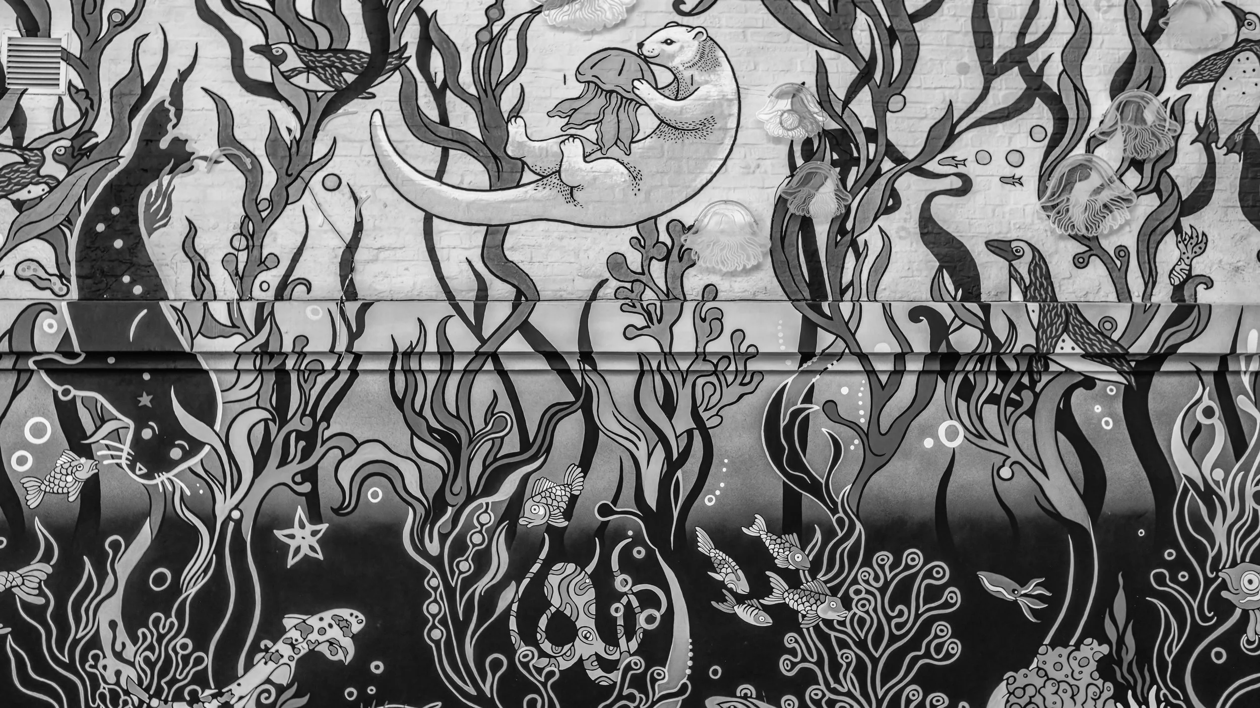 Black and white mural of an underwater scene with various fish, seaweed, and a large fish with a shell on its back, all on a brick wall background. Cape Town, South Africa.