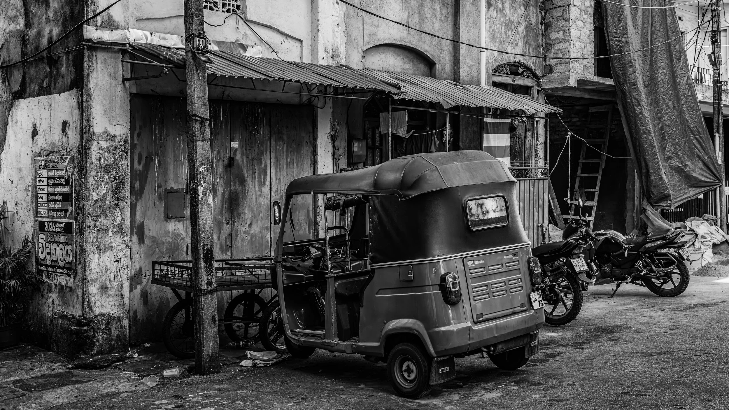 A black and white photo of an urban scene with a weathered wall, parked motorcycles, and a small three-wheeled vehicle in front, with string lights hanging above and construction materials on the right.