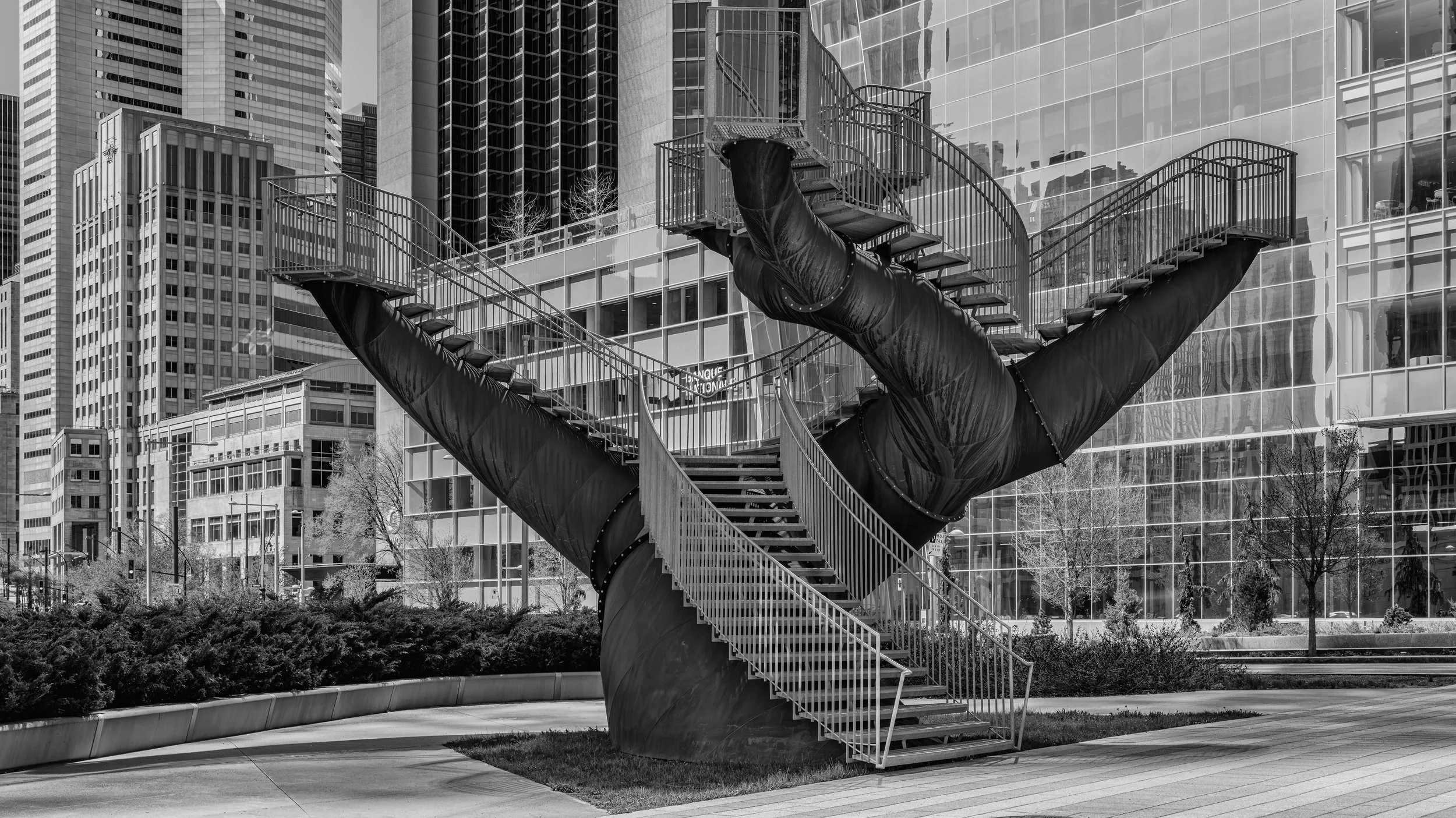 Modern urban scene with a large, unique sculpture resembling a pair of open hands, with spiral staircases in the palms, in front of tall glass office buildings.