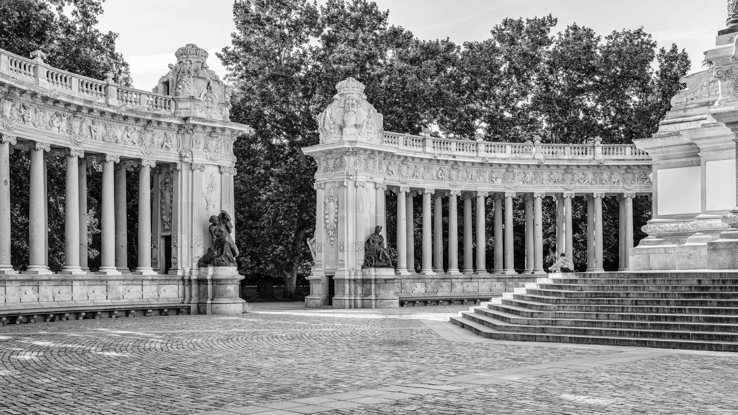 Black and white photo of an ornate classical-style stone structure with sculptures, columns, and steps, set amidst trees.