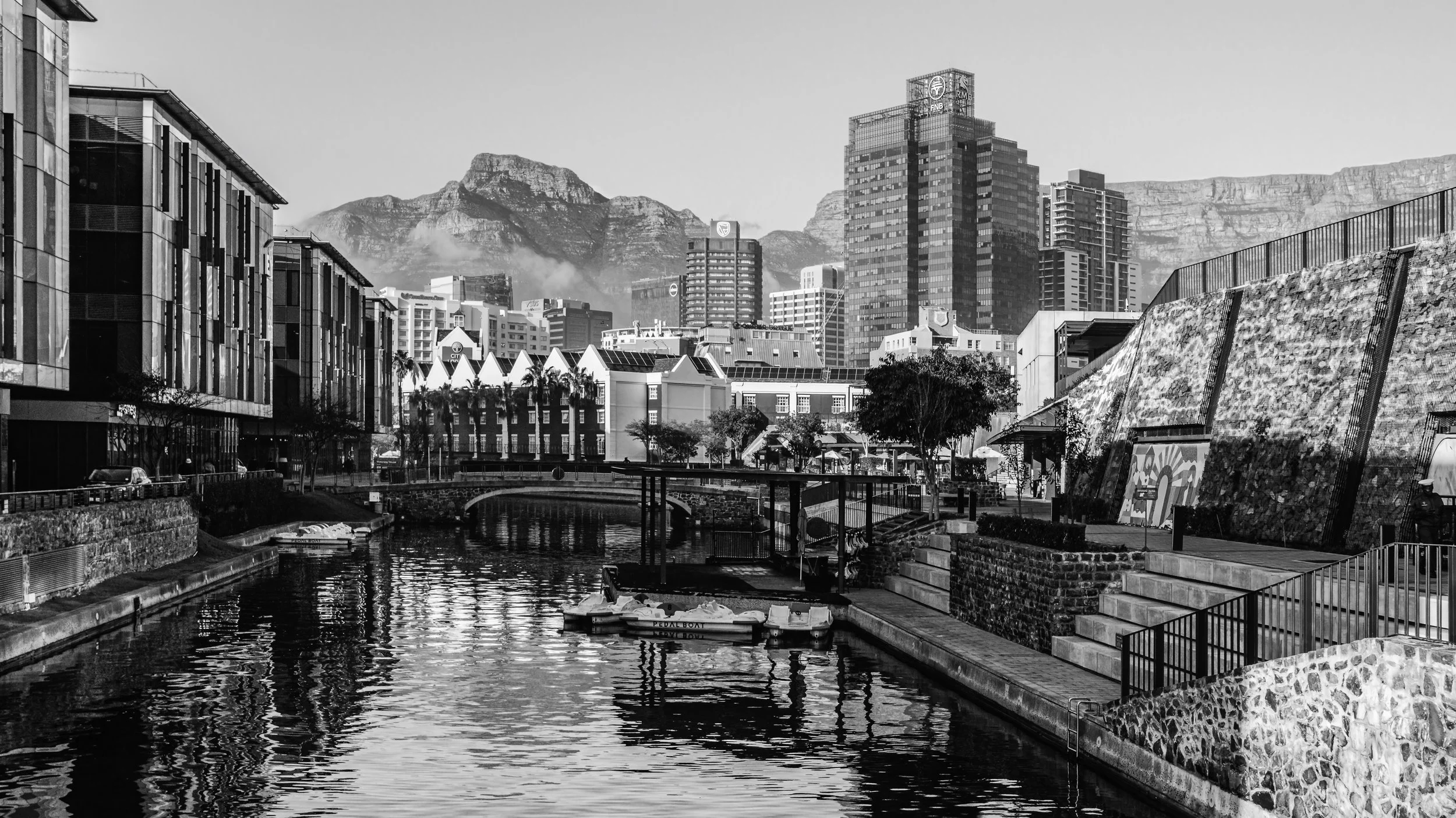 Black and white photo of a cityscape with modern buildings, mountains in the background, and a canal with a pedal boat in the foreground. Cape Town, South Africa.