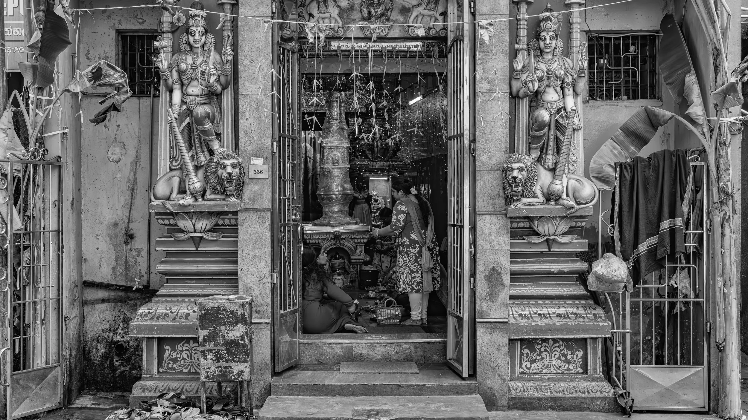 Black and white photo of a Hindu temple entrance with two large statues of female deities on either side, and intricate carvings. Several people are inside, with one woman standing and a woman sitting on the ground. Items like shoes and cloth are out