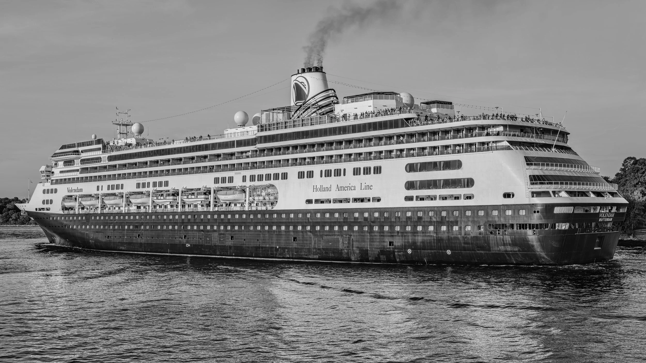 A large cruise ship named Volendam operated by Holland America Line sailing on water with trees in the background, black and white photograph.