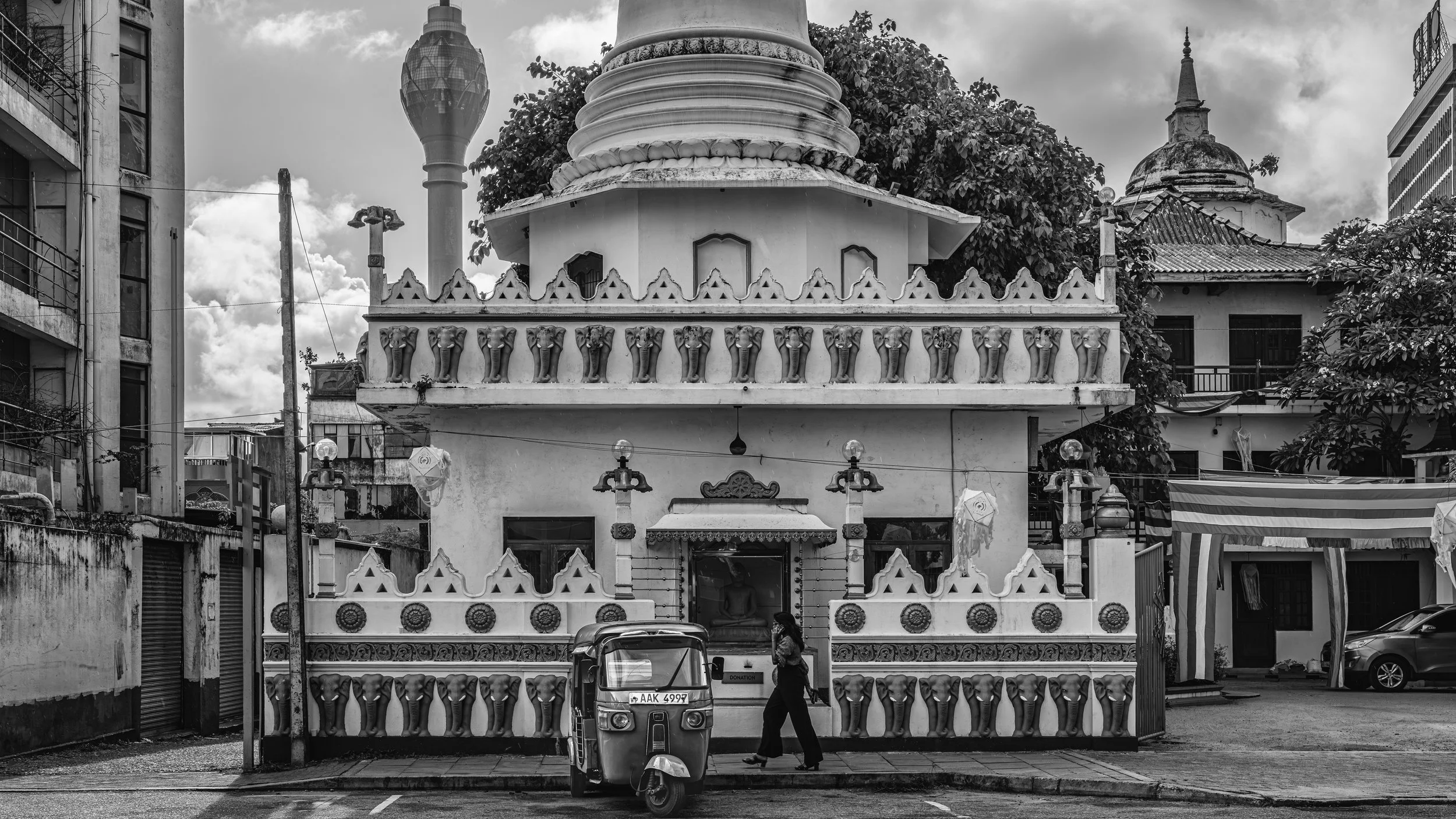 A black and white photo of a small temple or shrine with ornate decorations, elephant head motifs, and a dome in an urban setting. There are buildings, electric poles, a person walking, and a vehicle in front of the temple.