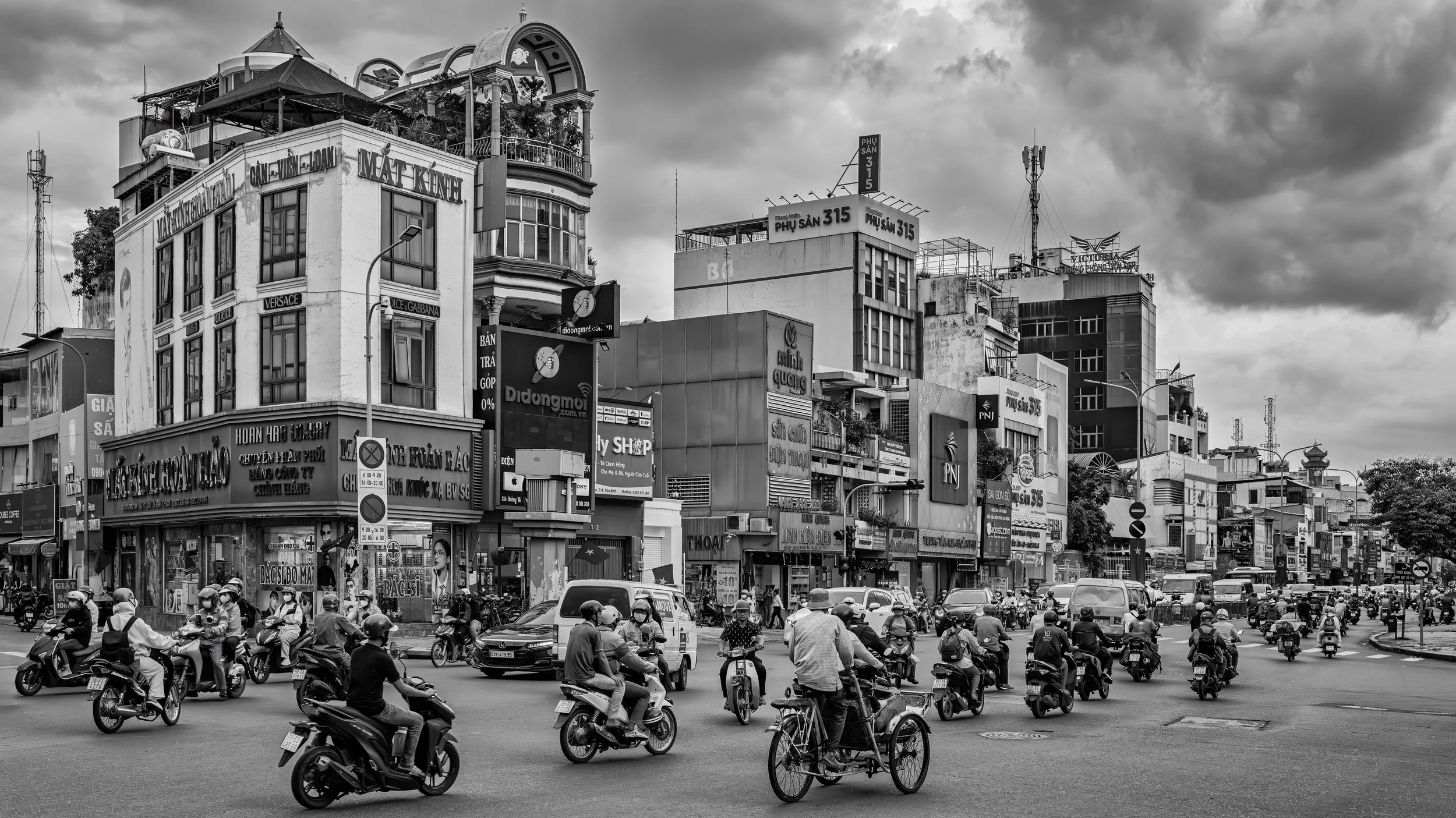 A busy city street filled with motorcycles and cars, with multi-story buildings and signage in the background under a cloudy sky.