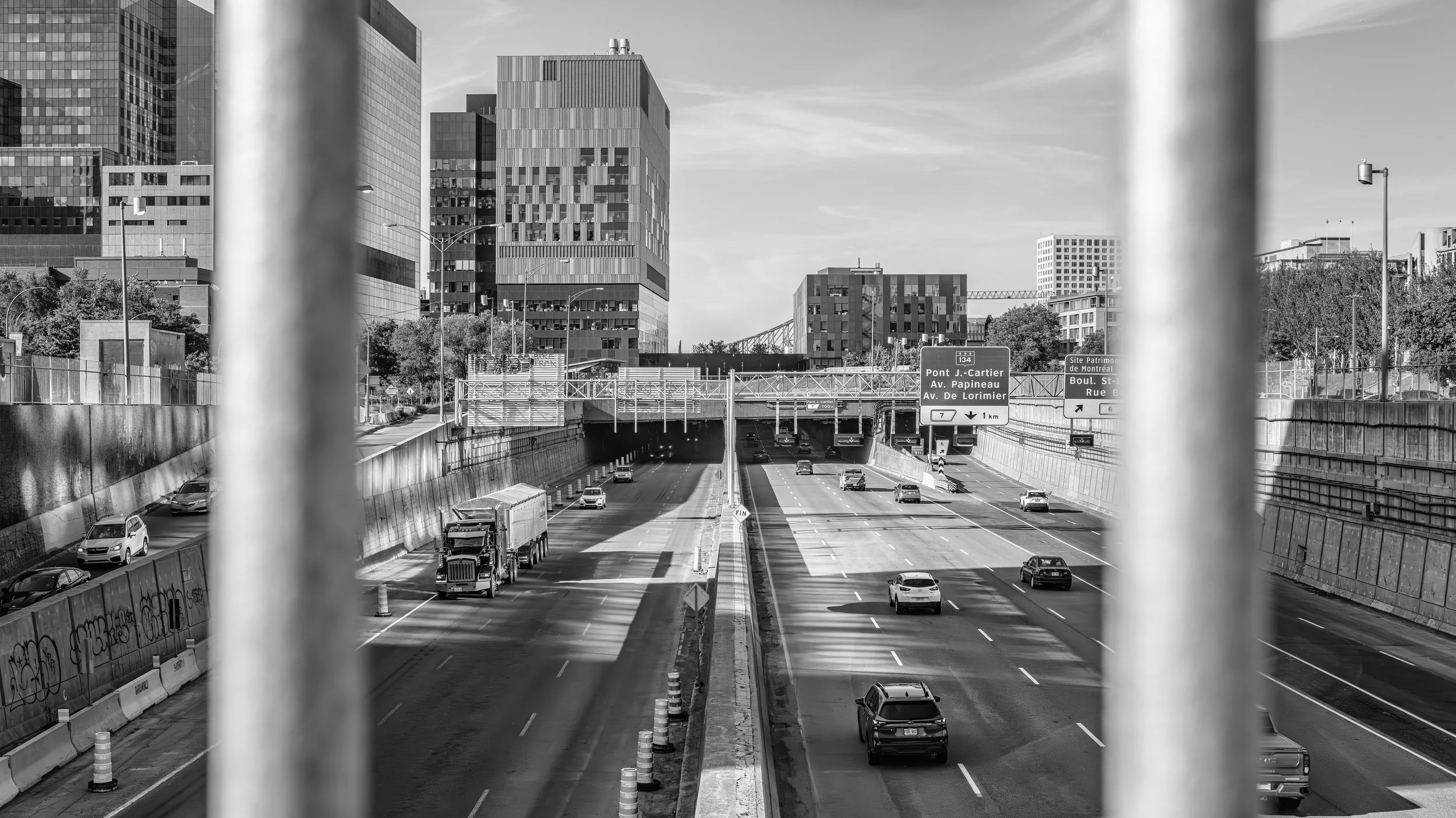 Black and white photo of a city highway with cars and trucks, overpasses, and modern tall buildings in the background, viewed through metal bars.