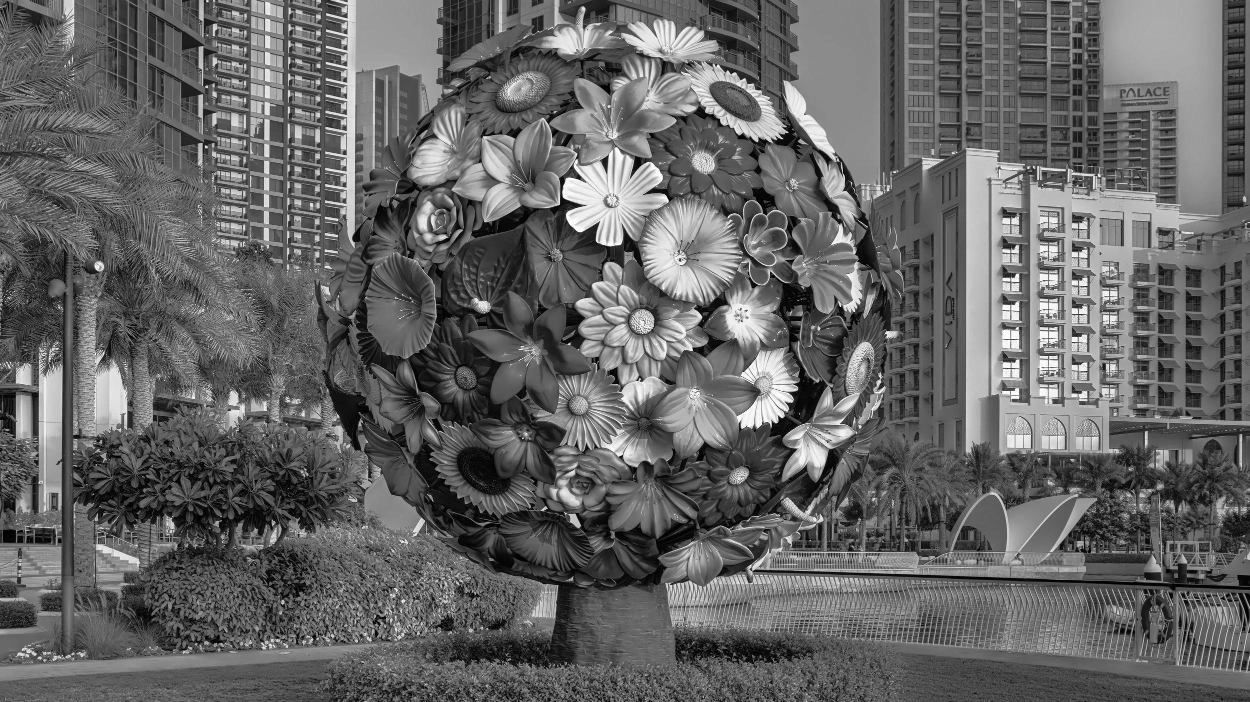 A large spherical sculpture made of numerous flowers in an urban park, with modern high-rise buildings in the background.