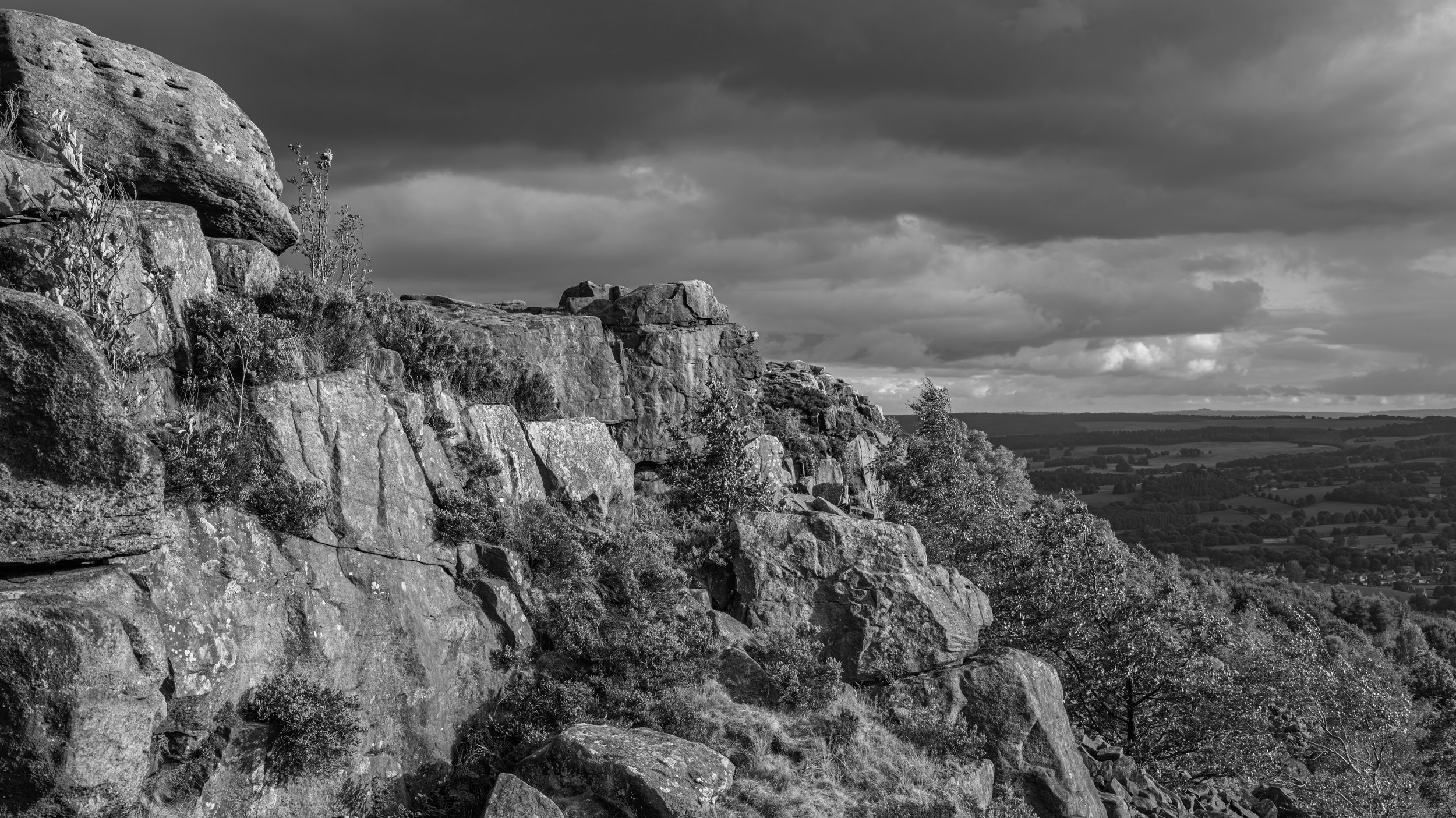 A black and white photo of a rocky hillside with large boulders and sparse vegetation, overlooking a vast landscape of fields and trees under a cloudy sky.