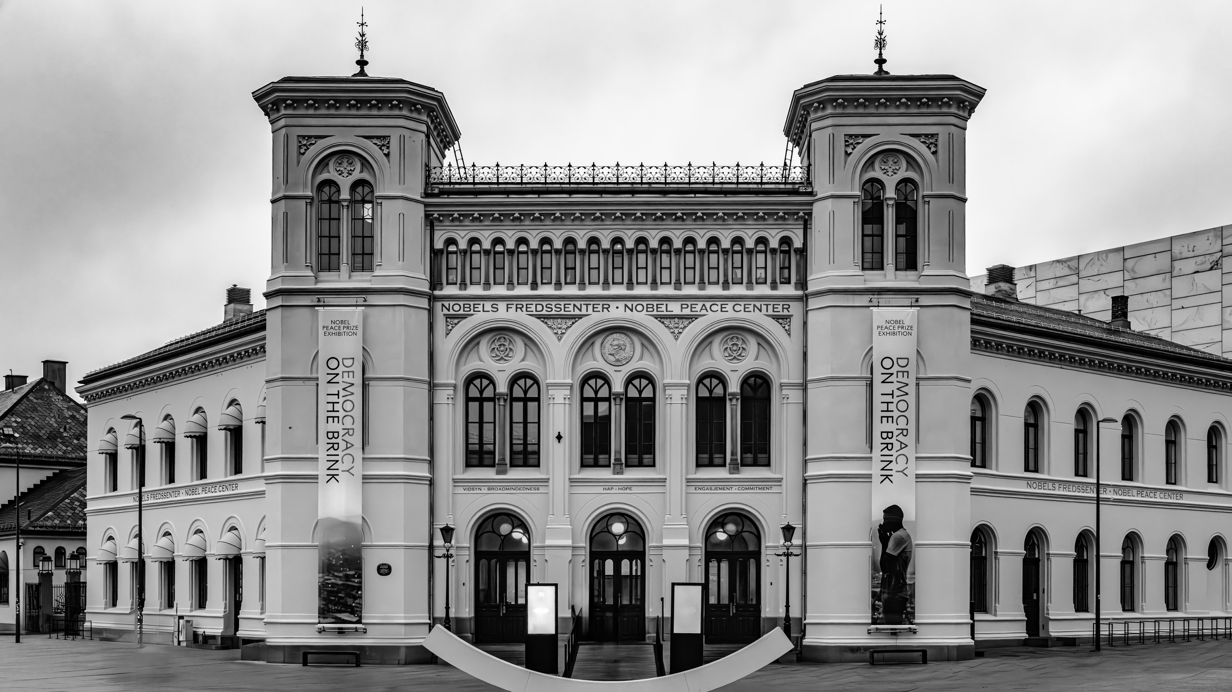 Black and white photograph of the Nobel Peace Center building in Oslo, Norway, with banners promoting an anti-war exhibition and messages about peace.