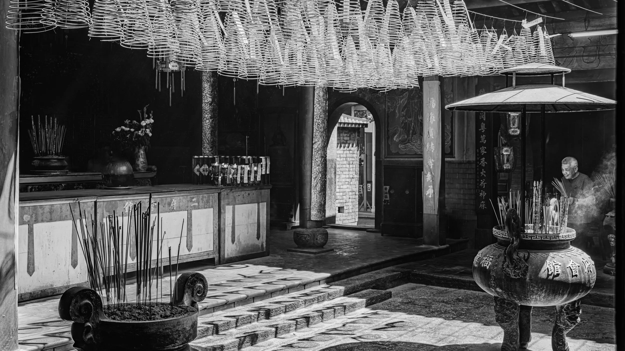 A black and white photo of a traditional Asian temple interior with incense burners, candles, and hanging lanterns.