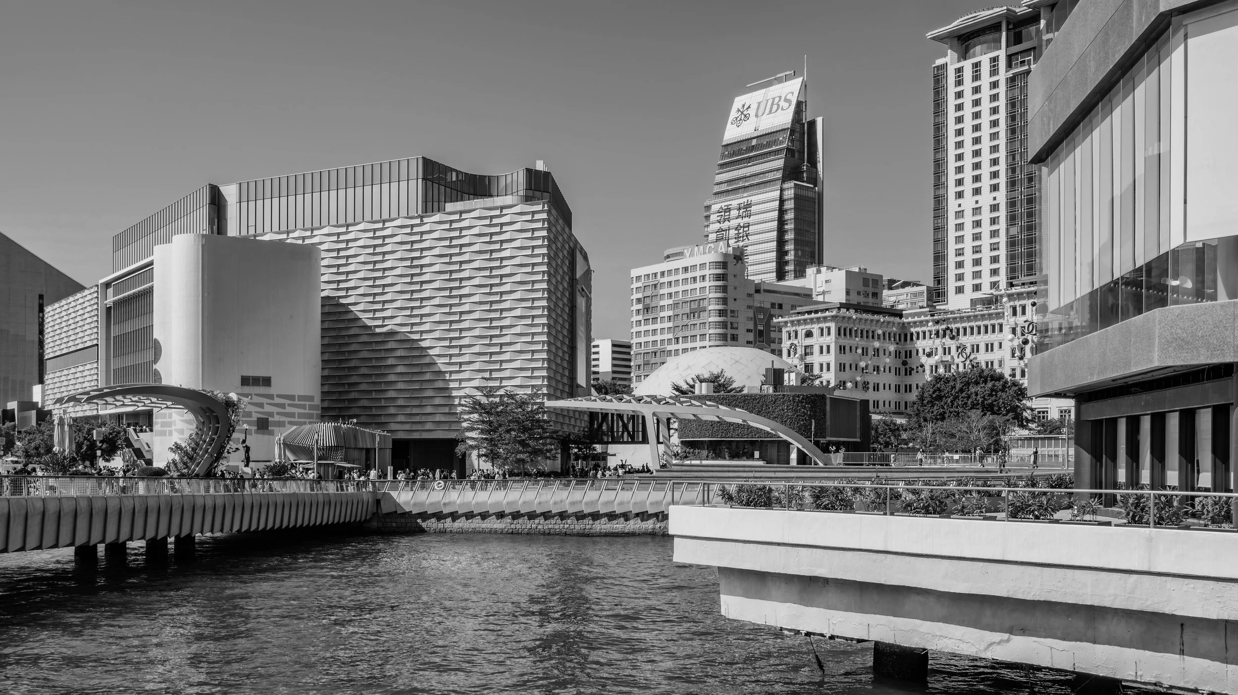 Black and white photo of a modern city skyline with tall buildings, a bridge over water, and a park area with trees and people.