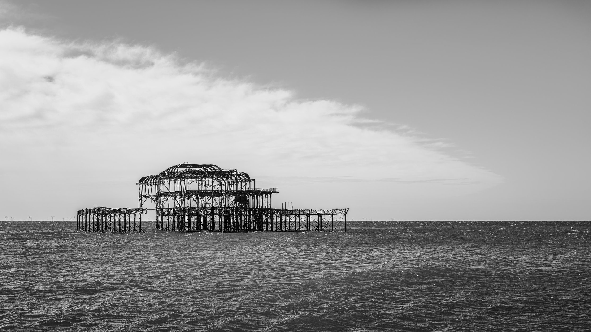 Black and white photo of a partially collapsed amusement park ride structure on the ocean with clouds in the sky.