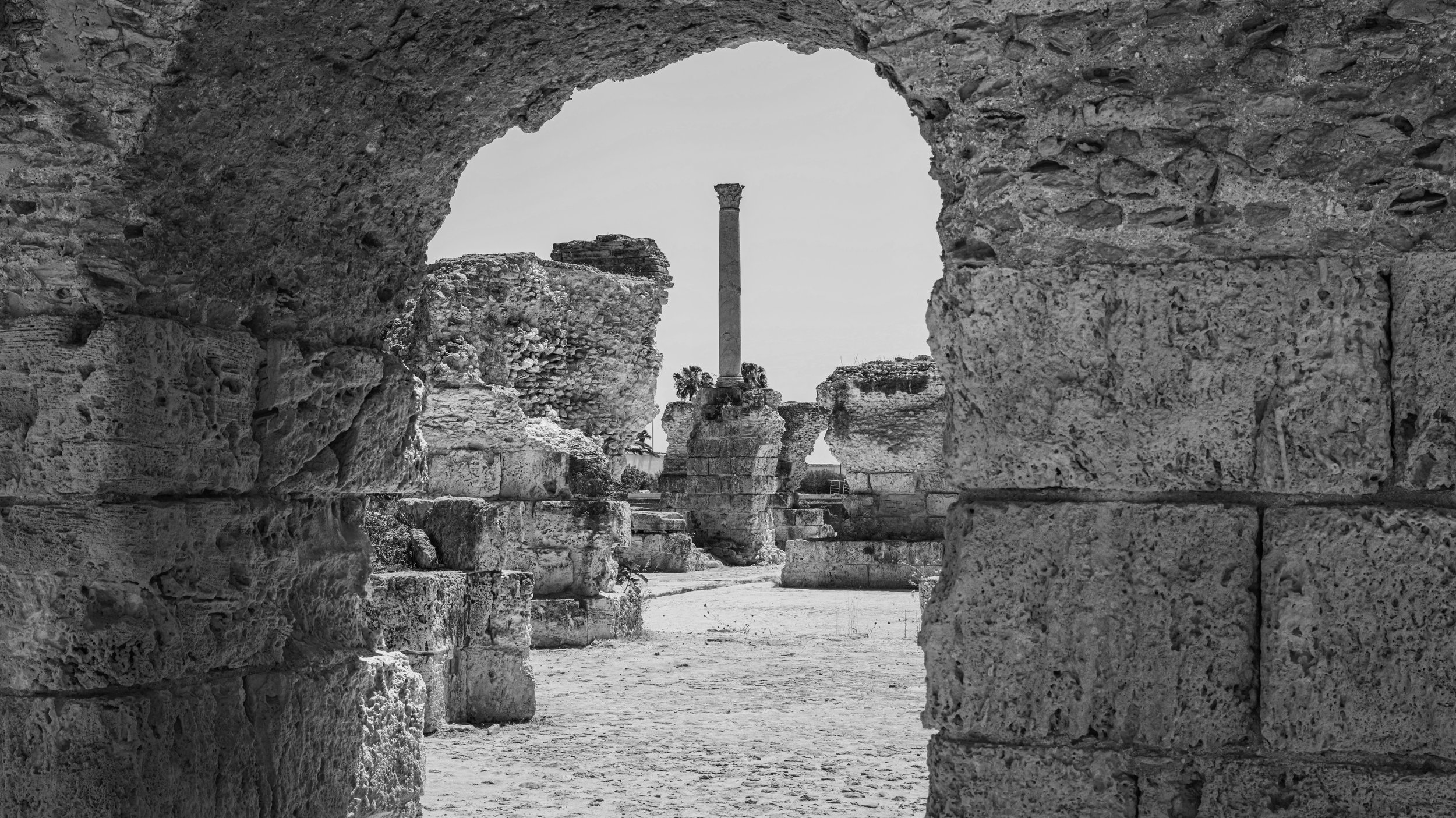 Black and white photograph of ancient ruins seen through a stone window or archway, with a tall column visible in the background.