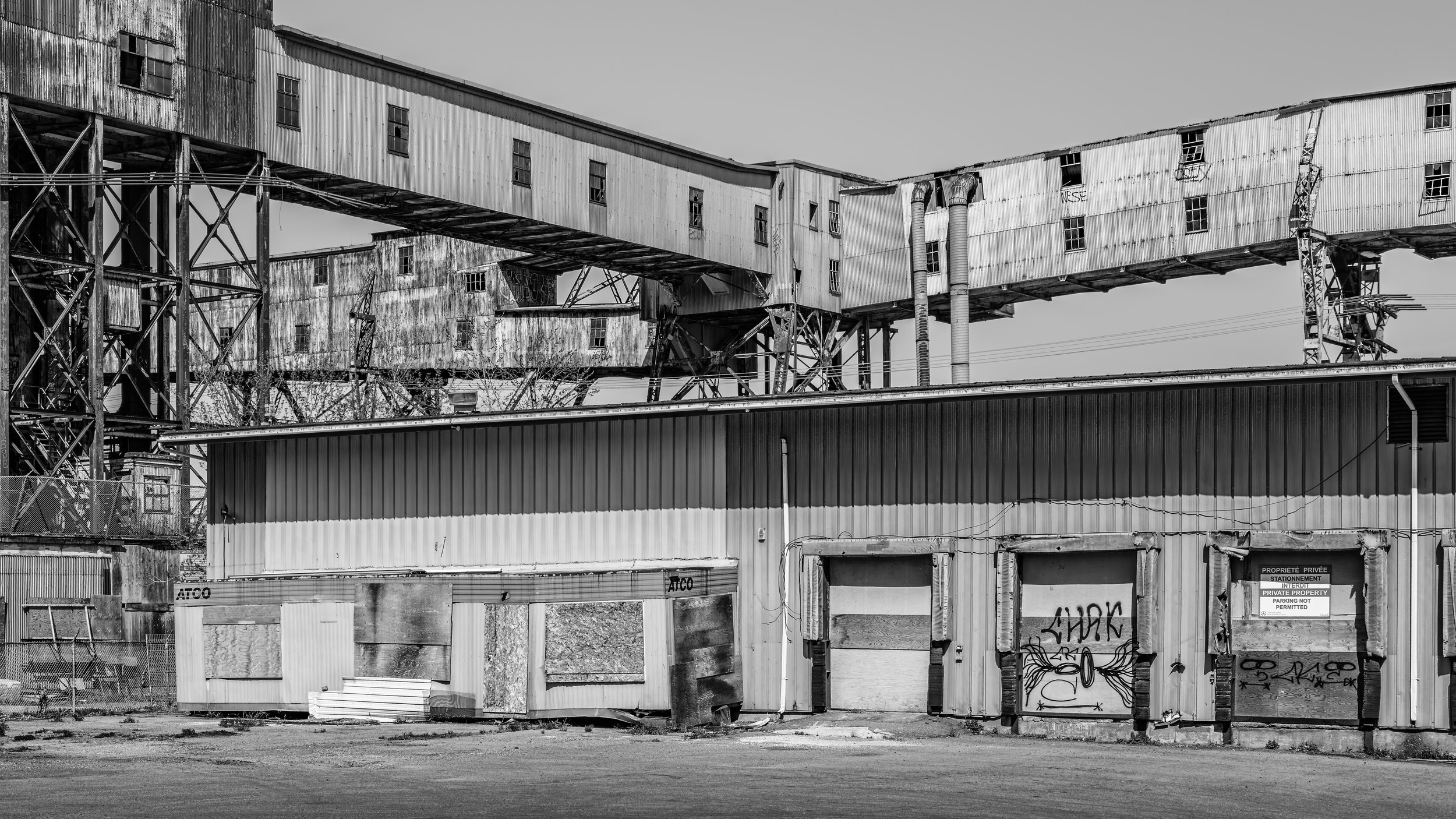 An abandoned industrial building with a deteriorated, rusted exterior and broken windows, with graffiti on walls, in black and white.