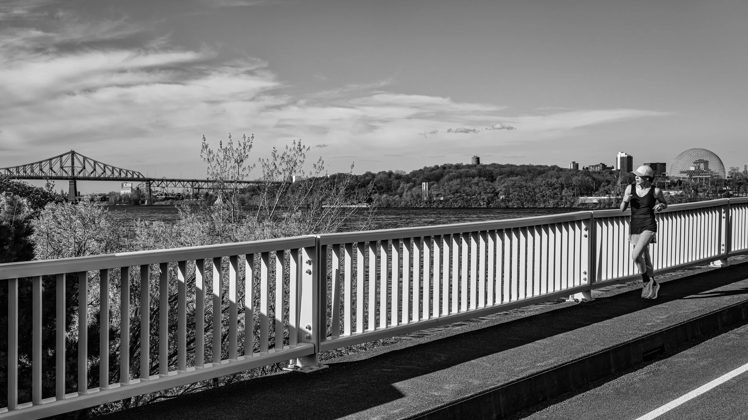 A woman jogs on a bridge near water with cityscape and a large geodesic dome building in the background in black and white.