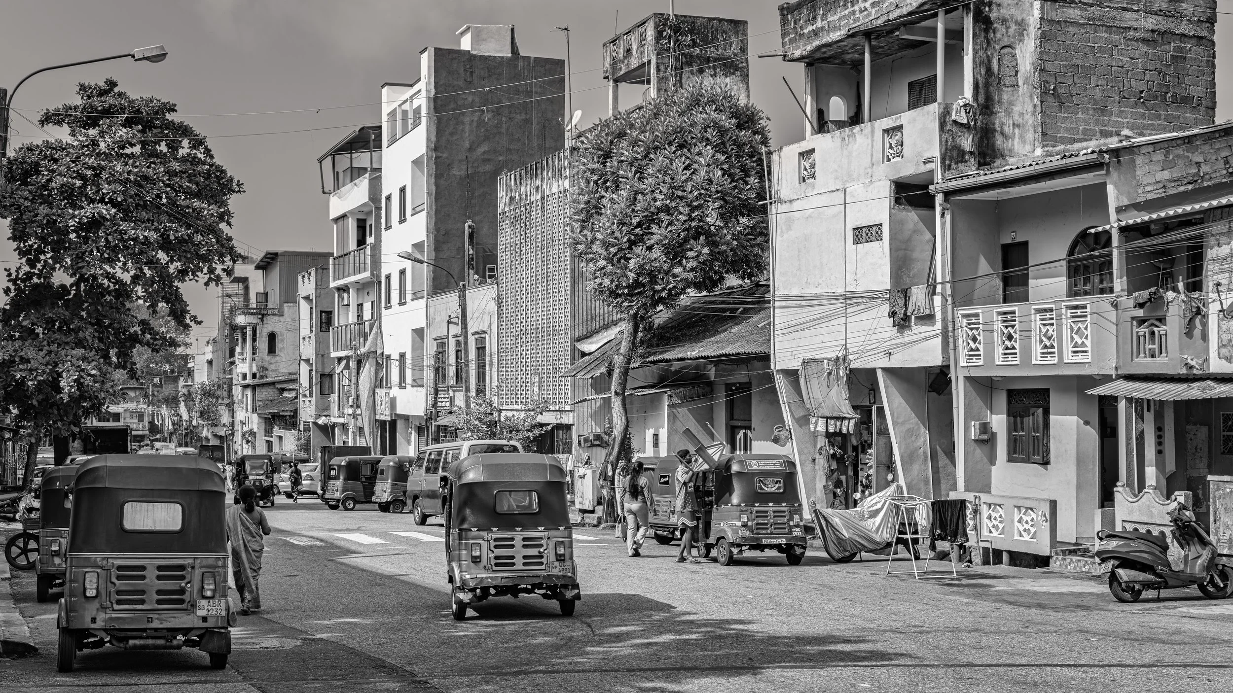 Street scene with parked auto rickshaws, pedestrians, and multi-story buildings in the background, some with laundry hanging and trees lining the street. Black-and-white photo.