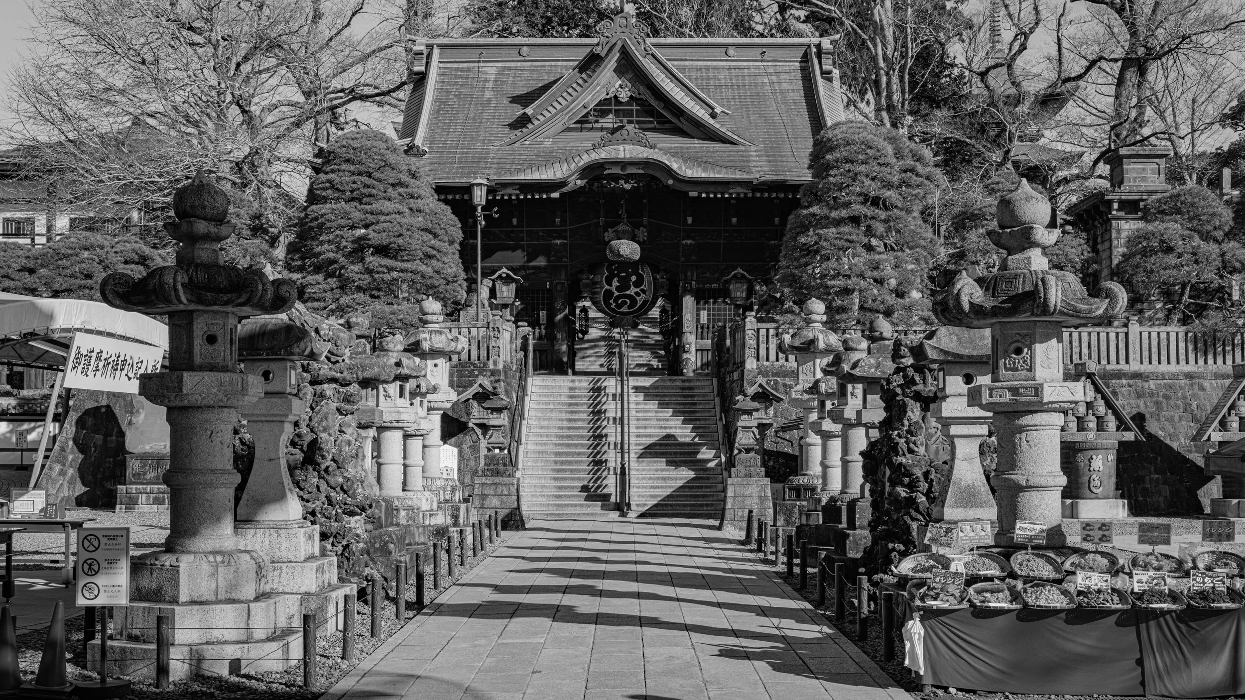 A traditional Japanese shrine with stone lanterns lining the stone steps leading to the shrine building. The shrine has a gabled roof and decorative wood elements, with trees in the background.